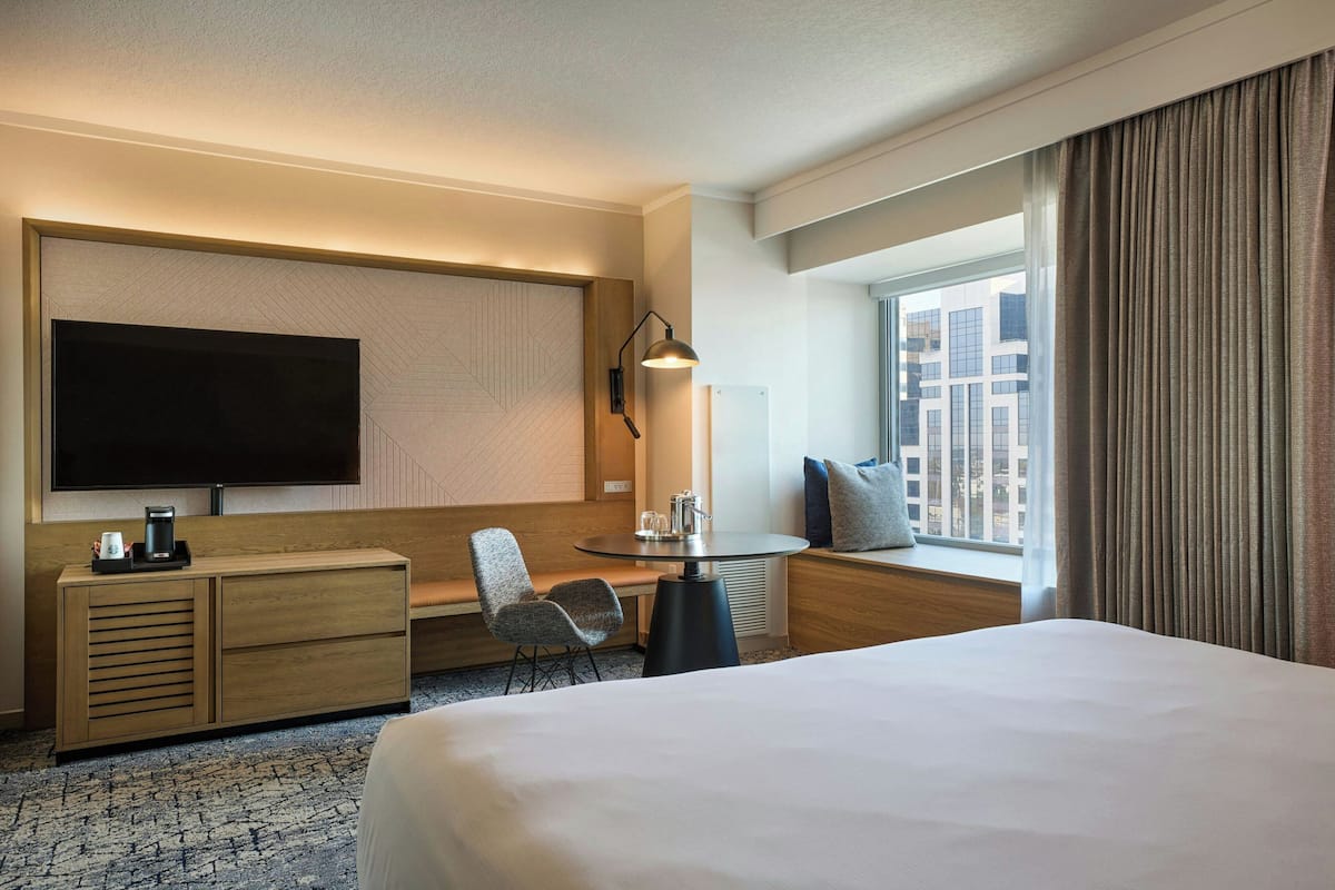 Ein Hotelzimmer mit einem großen Bett, einem Schreibtisch mit Stuhl, einem Flachbildfernseher, einer Kaffeemaschine und einem Fenster mit Vorhängen.