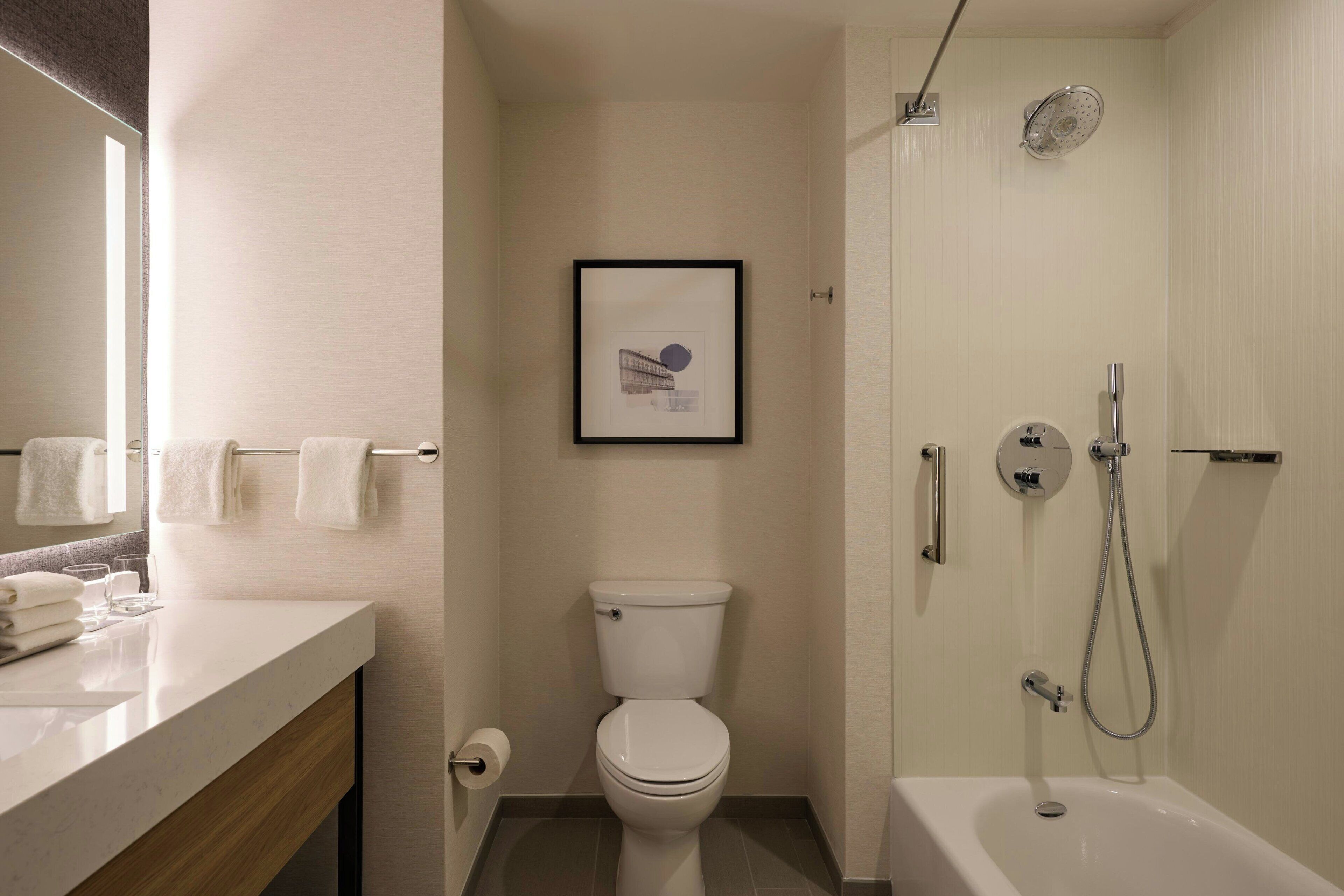 Et badeværelse med toilet, et billede i ramme på væggen, vask med håndklæder og et badekar med brusehoved.