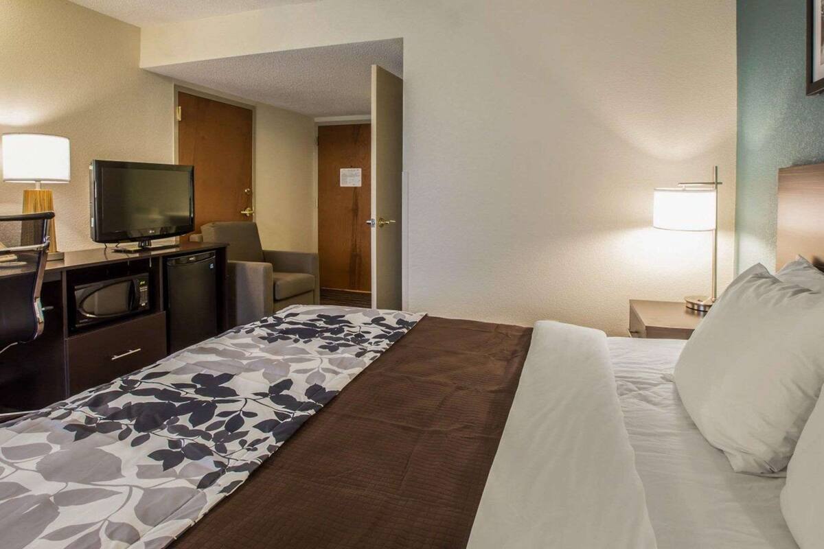 A hotel room with a bed, a desk with a television, a chair, and a bedside table with a lamp.