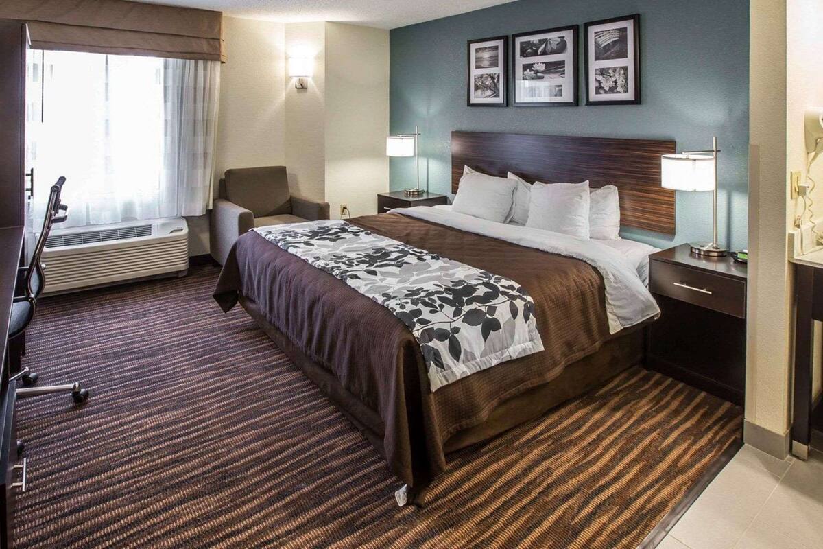 A hotel room with a large bed, a desk, a chair, a desk lamp, a wall-mounted TV, and a window with curtains.