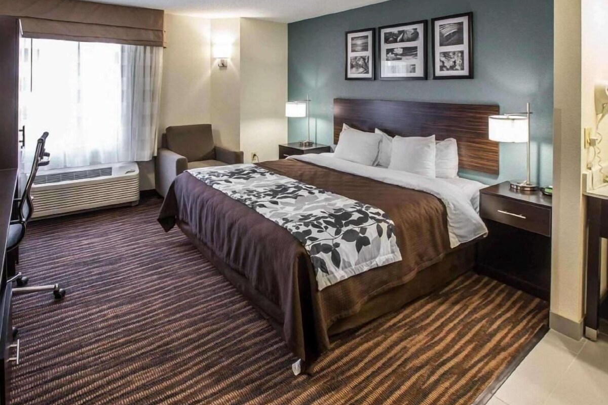 A hotel room with a bed, bedside tables, a chair, a desk, a lamp, a picture frame, and a window with curtains.