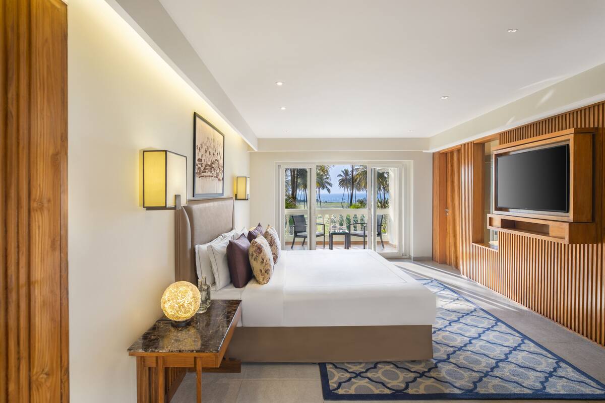 Habitación de hotel con cama, televisor, balcón y vistas a palmeras, además de paneles de pared de madera.
