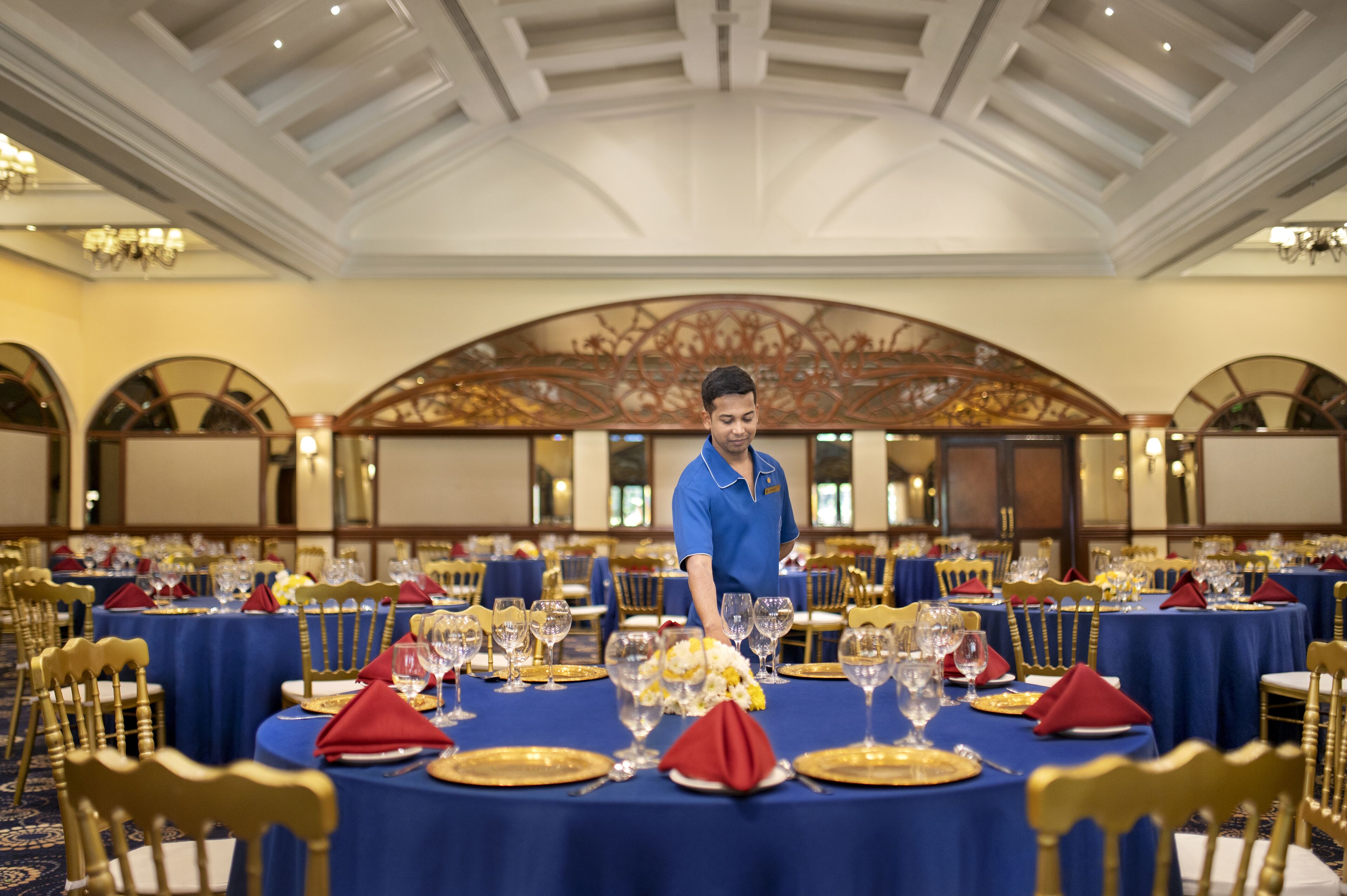 A banquet hall with round tables set for a formal event, a waiter in uniform, and ornate ceiling architecture.