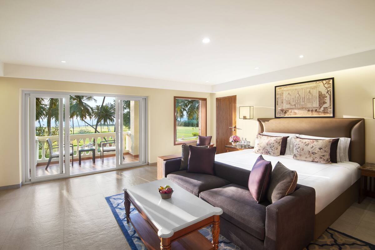 Un dormitorio amplio con una cama grande, un sofá, una mesita y un balcón con vistas a palmeras.