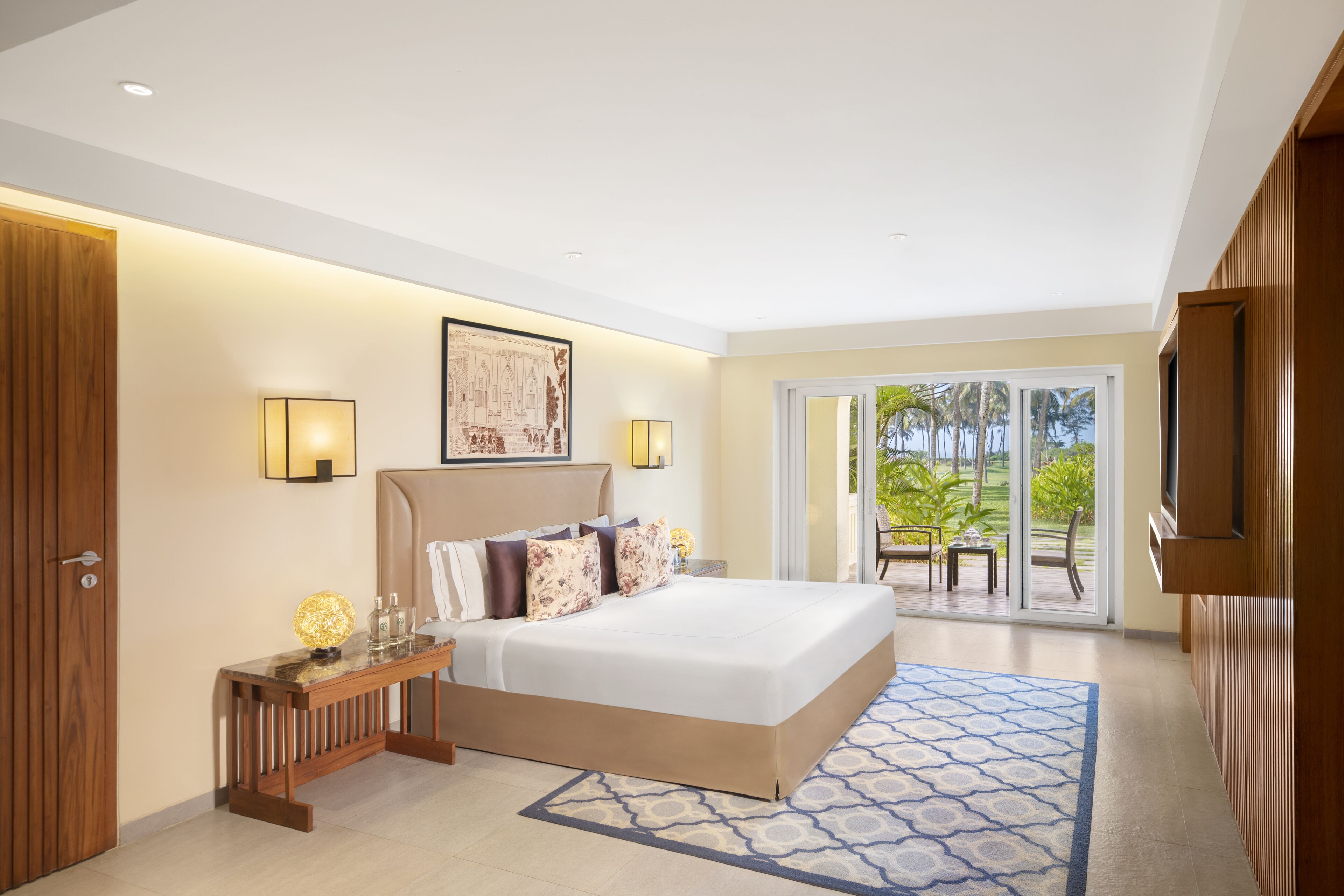 A hotel room with a large bed, a wooden nightstand, a wall-mounted TV, and a balcony with outdoor seating.