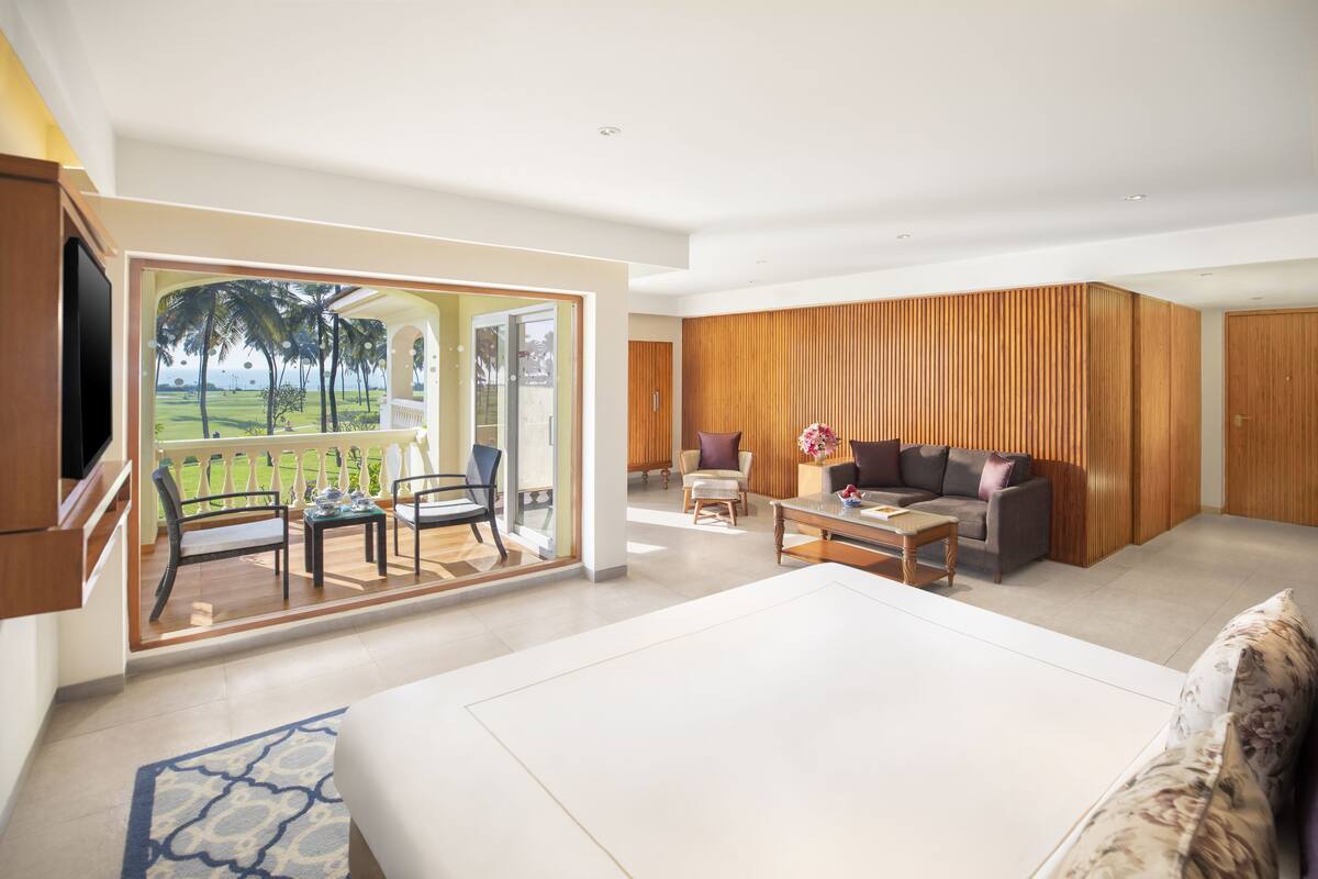 Habitación de hotel moderna con una cama grande, un balcón con vistas a palmeras, una zona de estar con sofá y sillones, y paneles de madera en las paredes.