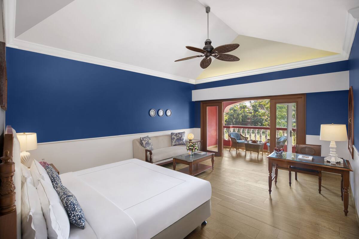 Habitación de hotel con una cama grande, ventilador de techo, sofá, una mesa de centro y una mesa de comedor. Cuenta con un balcón con asientos exteriores.