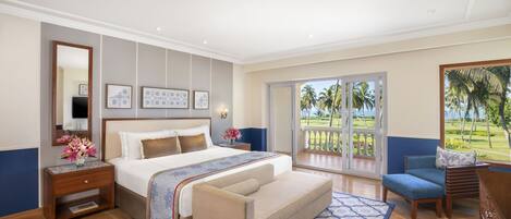 Habitación de hotel con una cama grande, un área de descanso con sofá y sillón, una mesita de noche de madera, un espejo y vistas a palmeras a través de una puerta corrediza de vidrio.