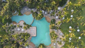 Vista aérea de un resort con una piscina central, rodeado de palmeras y sillones de sol.