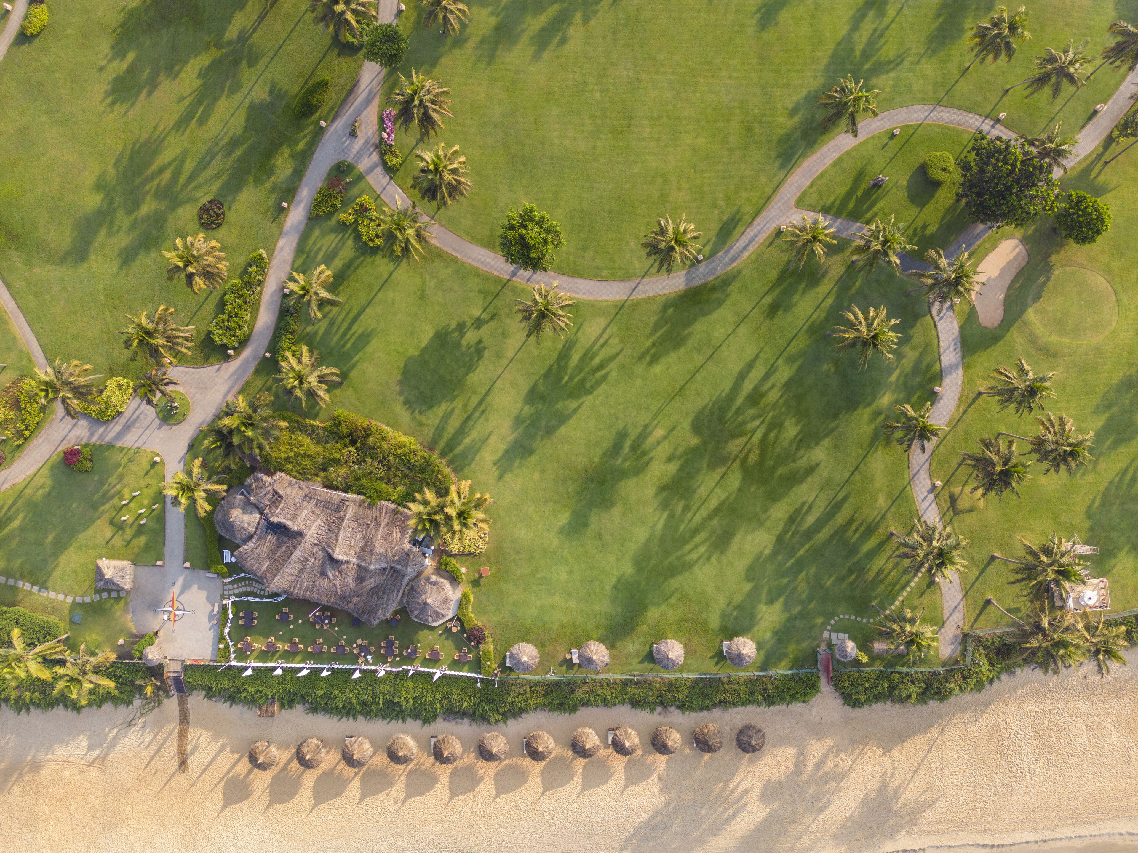 Luftaufnahme eines Resorts mit einer zentralen Felsformation, Palmen und einem Strandabschnitt mit Sonnenschirmen.