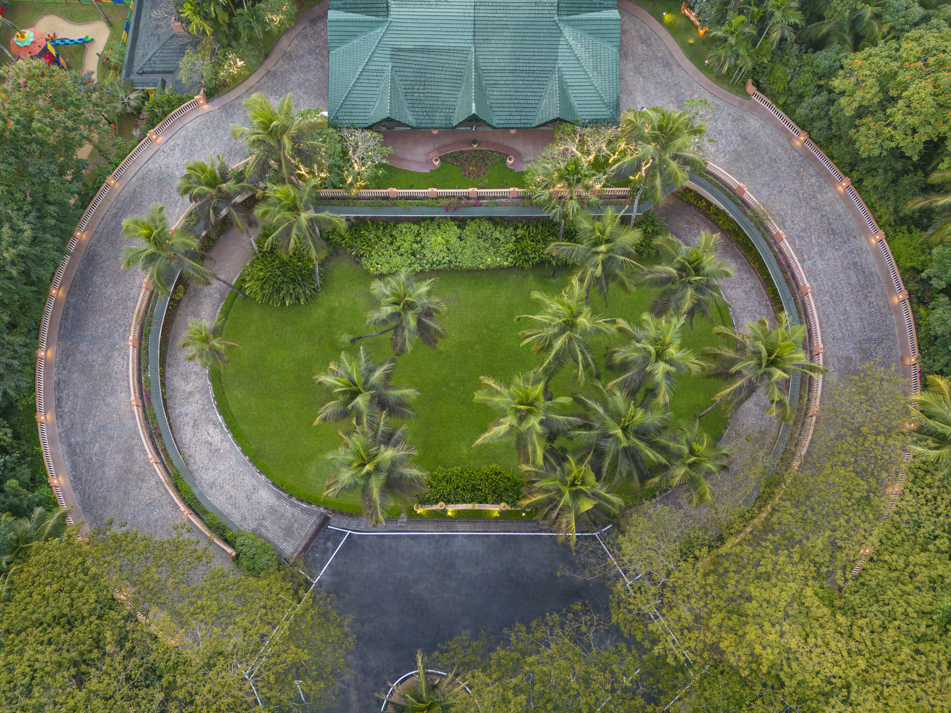 An aerial view of a circular park with palm trees, a central lawn, and a building with a green roof.