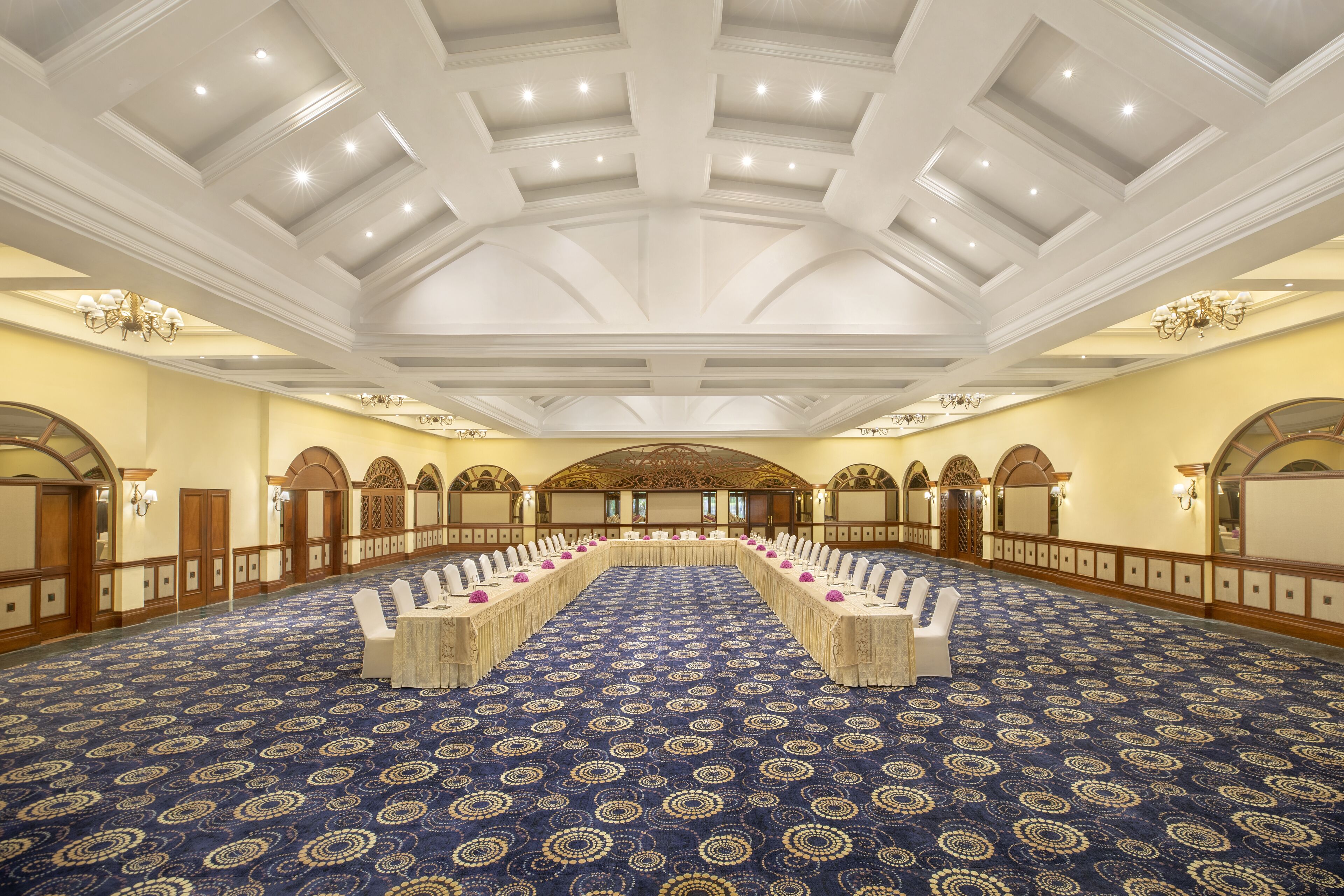 A large conference room with a patterned carpet, white tables, and chairs, and a high ceiling with recessed lighting.