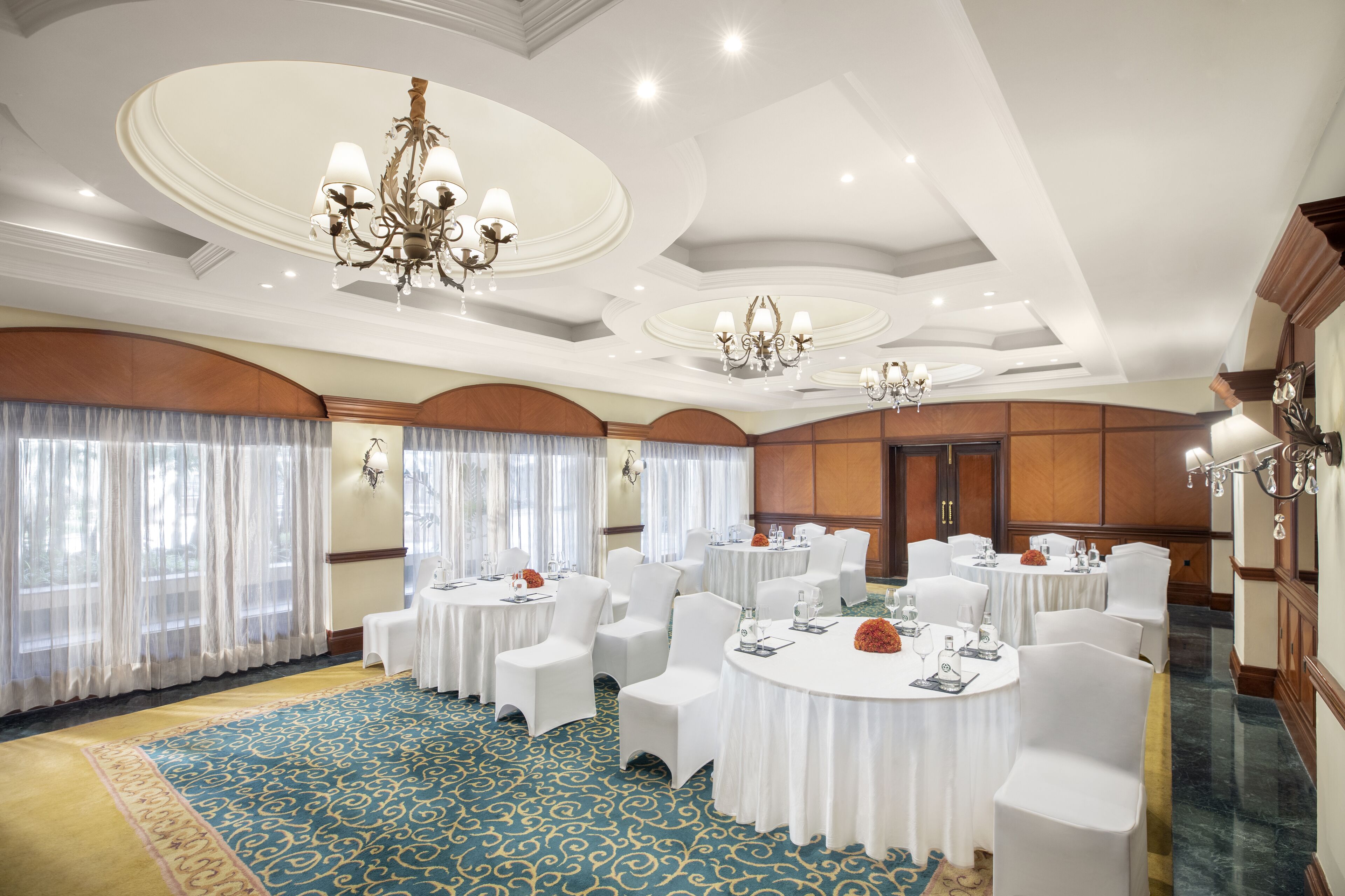 Un salón de banquetes con mesas redondas, sillas blancas, candelabros y una alfombra azul estampada.