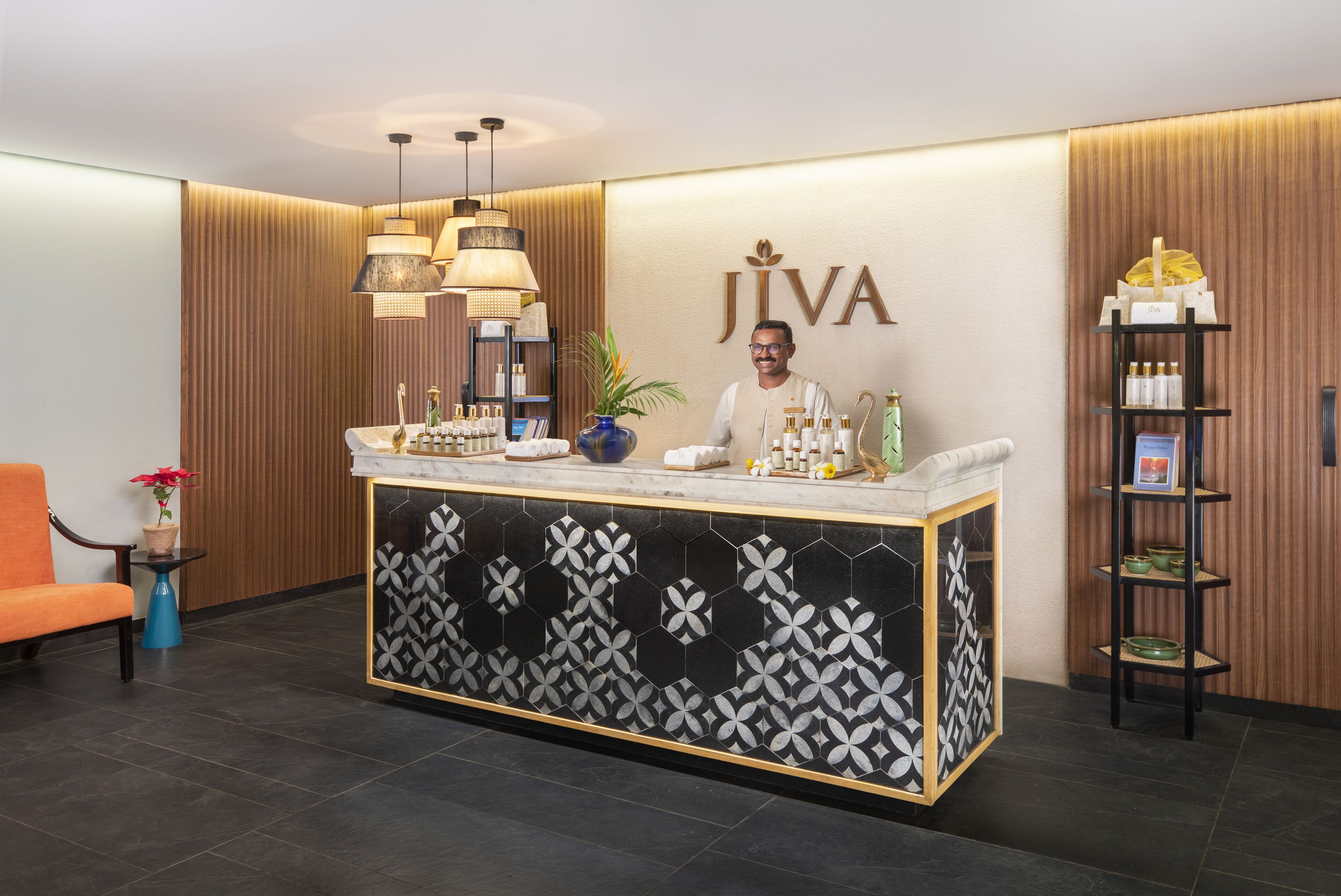 A reception desk with a logo, a man standing behind it, and a shelf with bottles and a vase.