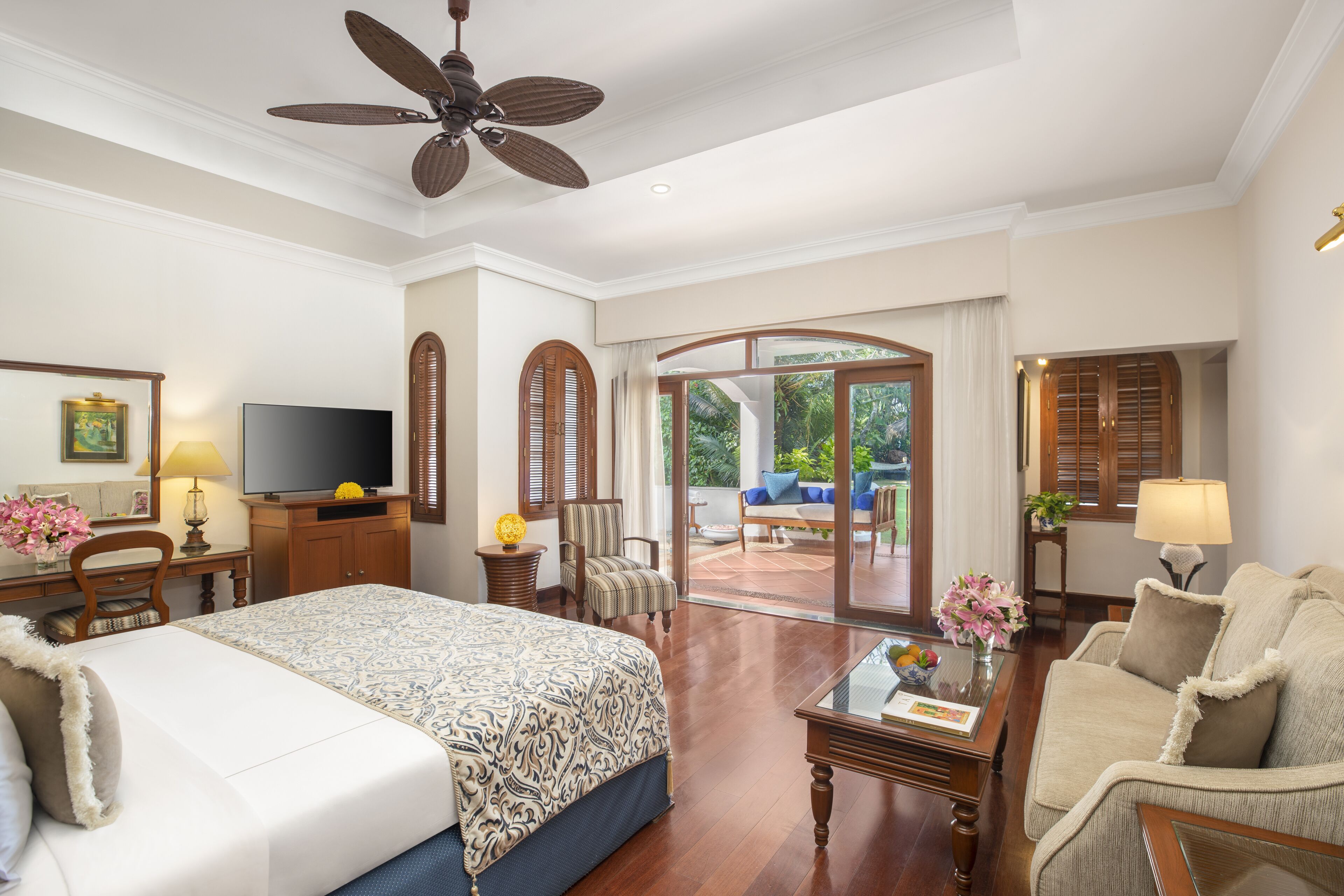 A hotel room with a large bed, a television, a desk with a lamp, a chair, a sofa, and a sliding glass door leading to an outdoor area.