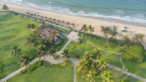 Vista aérea de un complejo turístico en la playa con una estructura de techo de paja, palmeras y un camino que conduce a la playa.