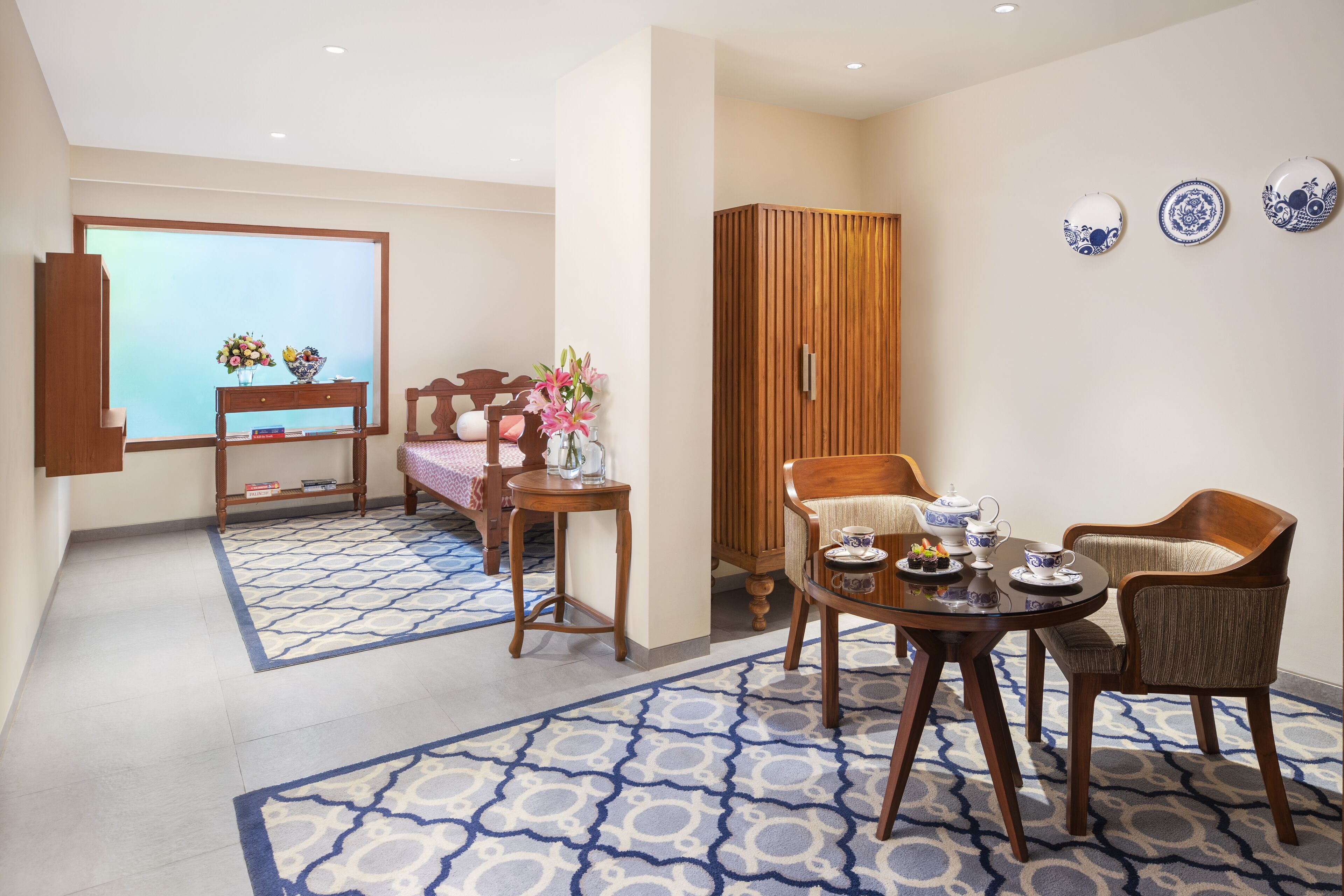 A hotel room with a large window, a wooden wardrobe, a dining table with chairs, and a small side table with a vase of flowers.