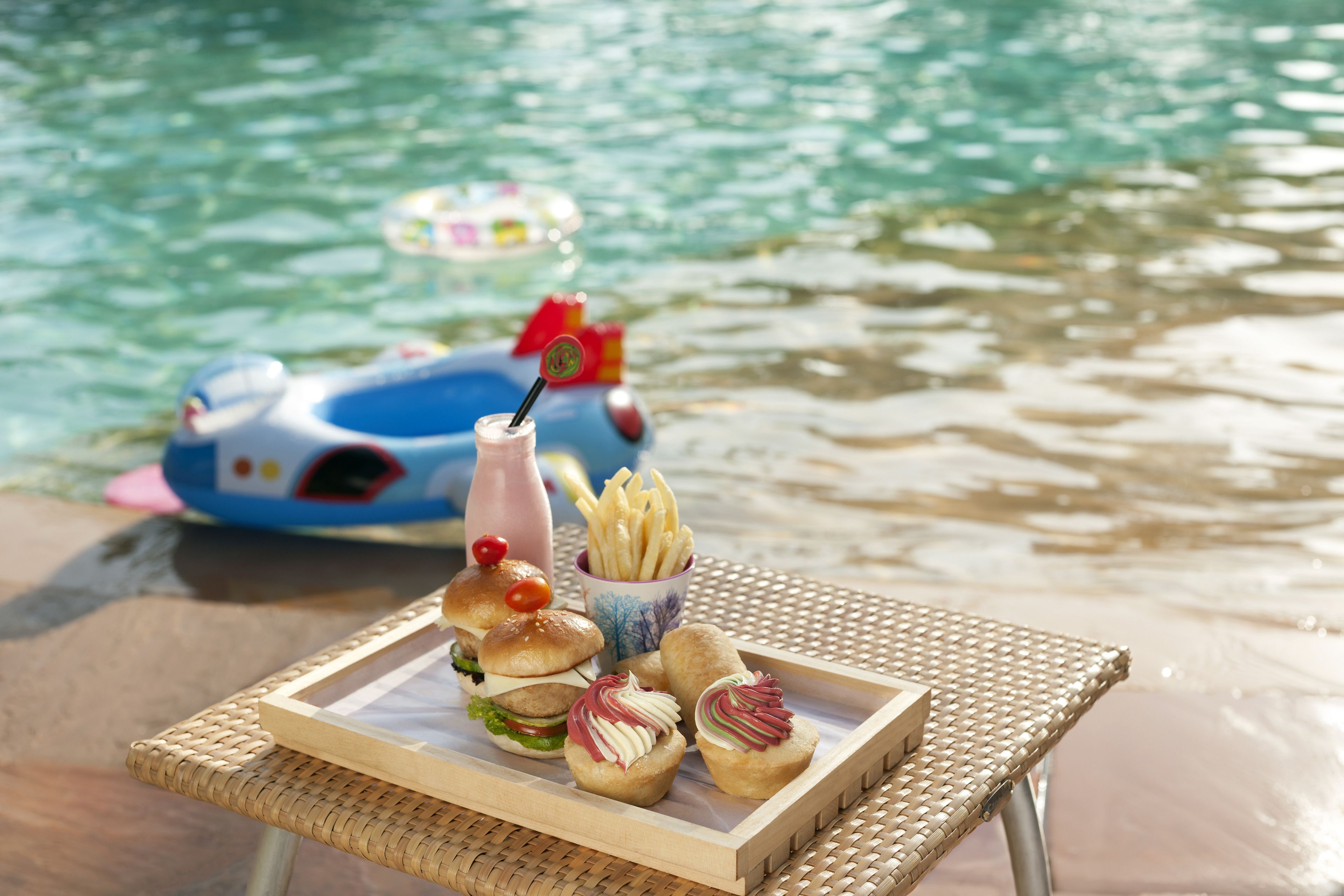 A wicker table with a tray of food including burgers, fries, and cupcakes, near a pool with a blue float.