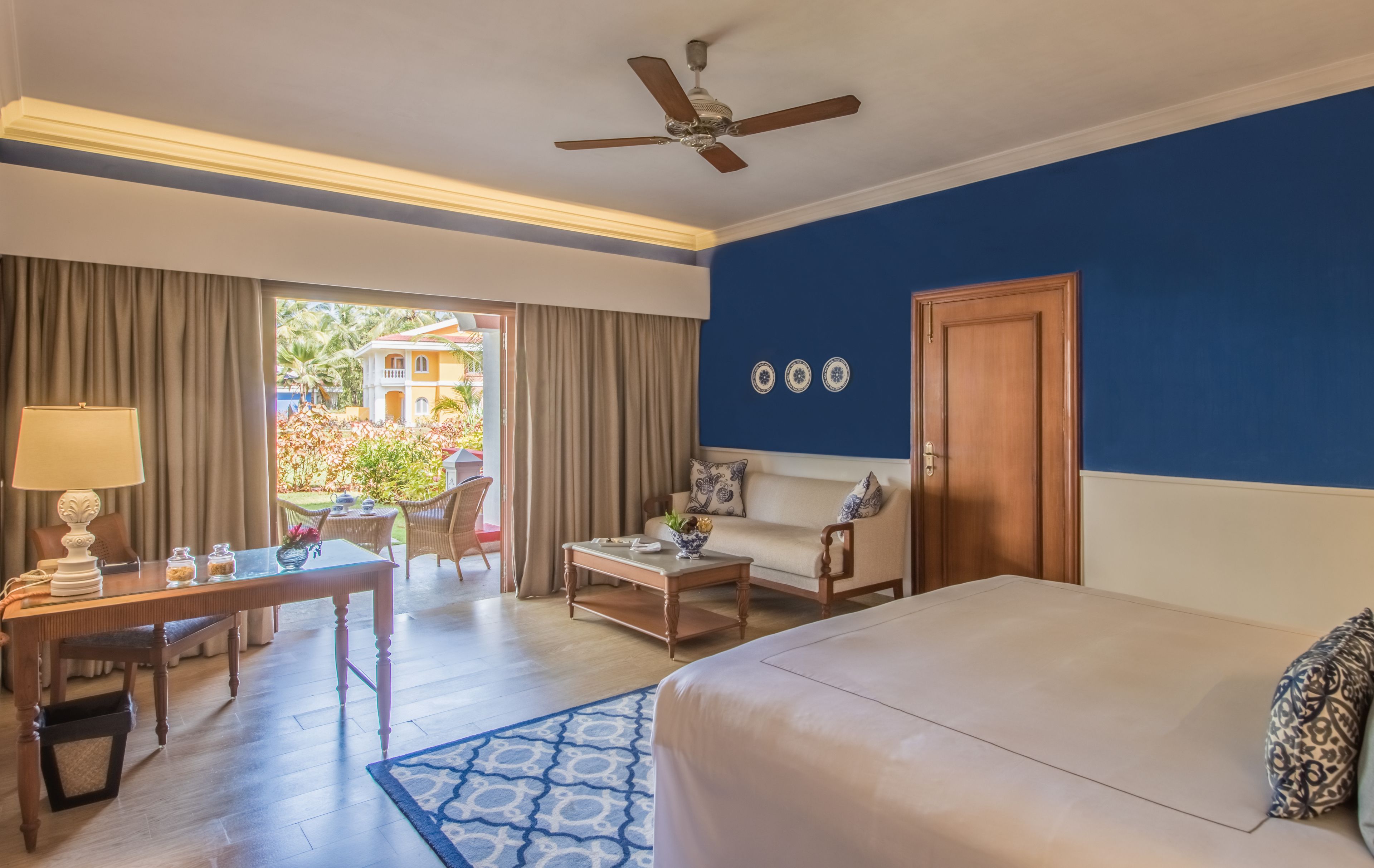 A hotel room with a bed, a desk with a lamp, a coffee table, a sofa, and a view of the outside.