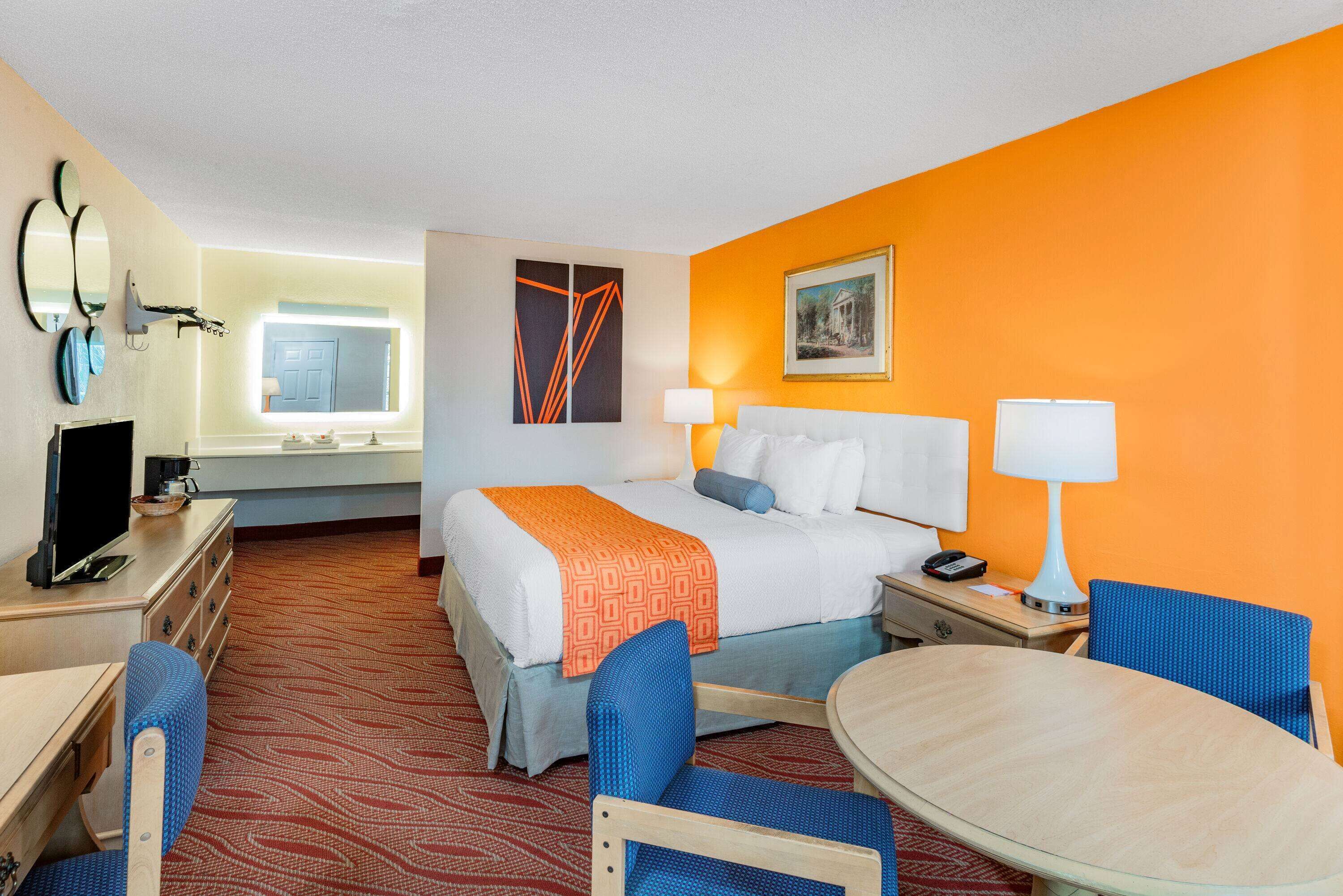 A hotel room with a bed, a desk, a chair, a table, and a lamp.