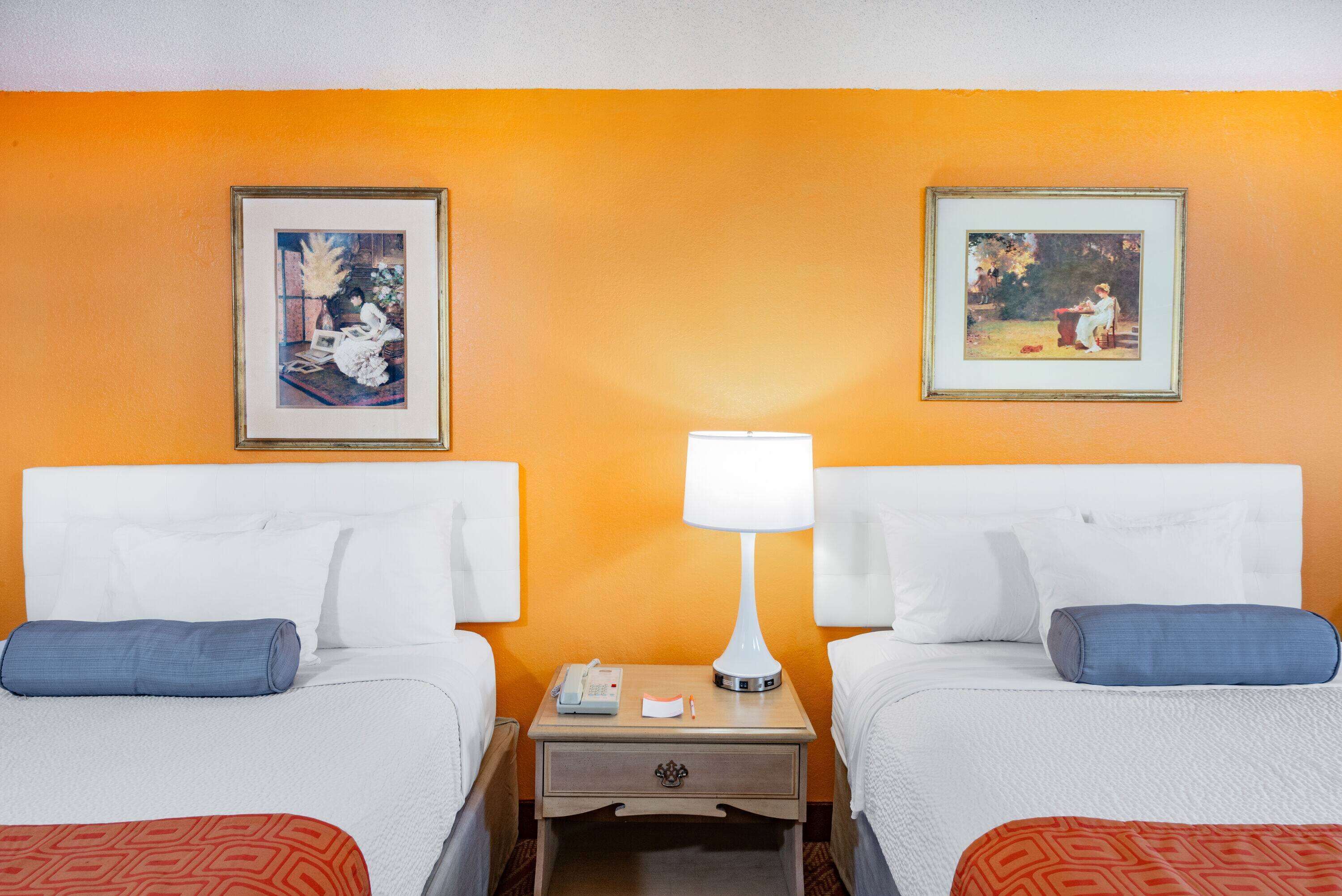 Habitación de hotel con dos camas, una mesita de noche con lámpara y dos cuadros en la pared.