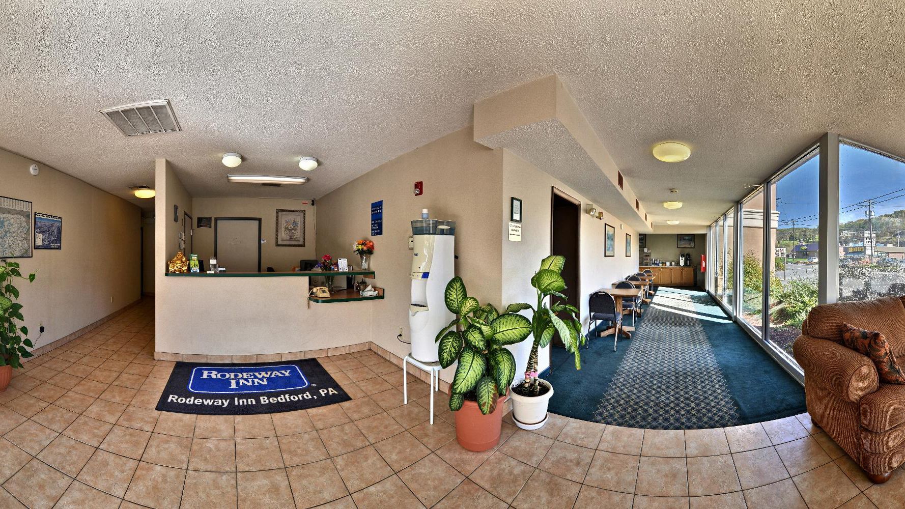 A hotel lobby with a reception desk, potted plants, a dining area, and large windows offering a view.