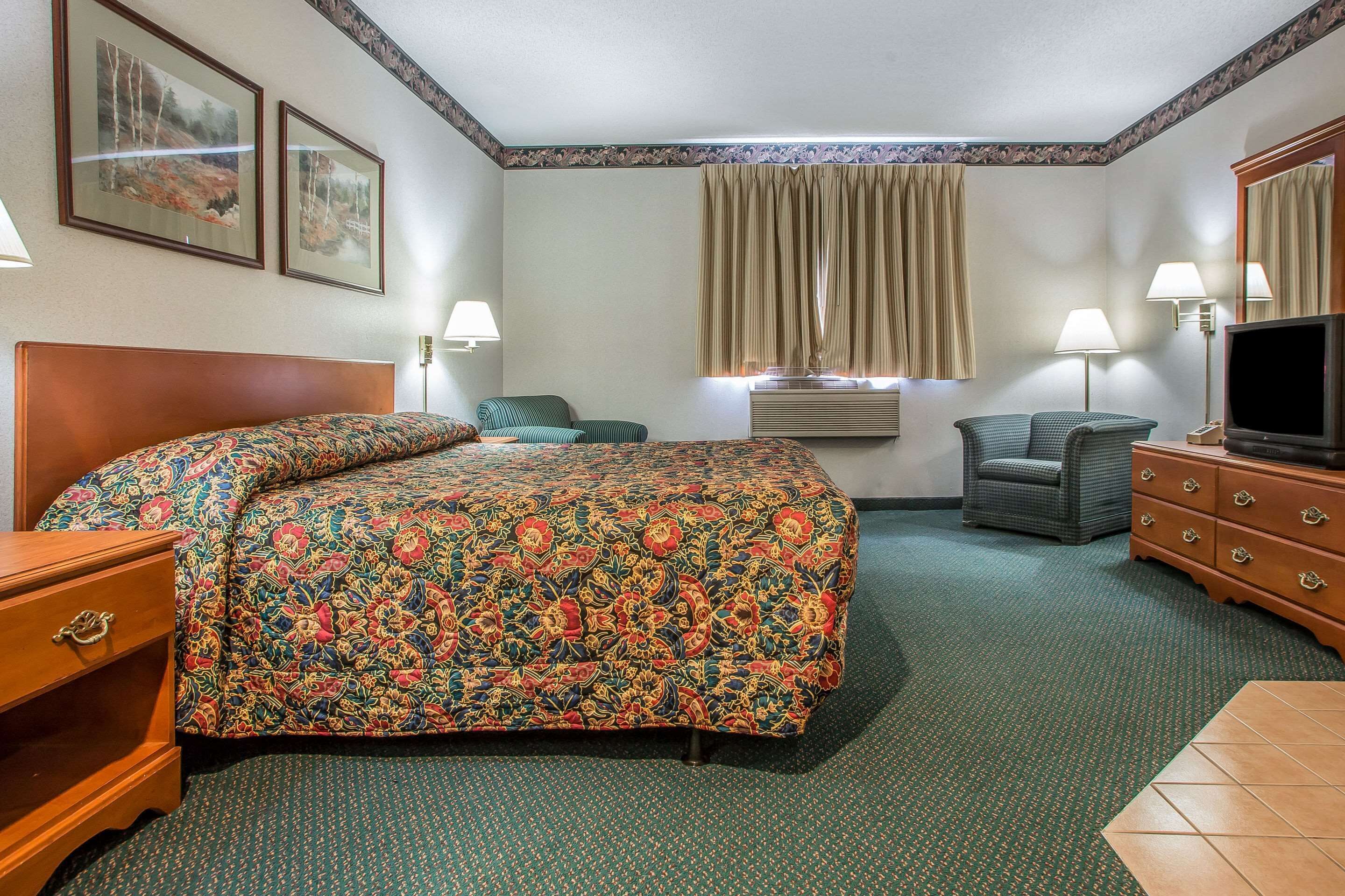 A hotel room with a large bed, two chairs, a dresser, a television, and framed pictures on the wall.