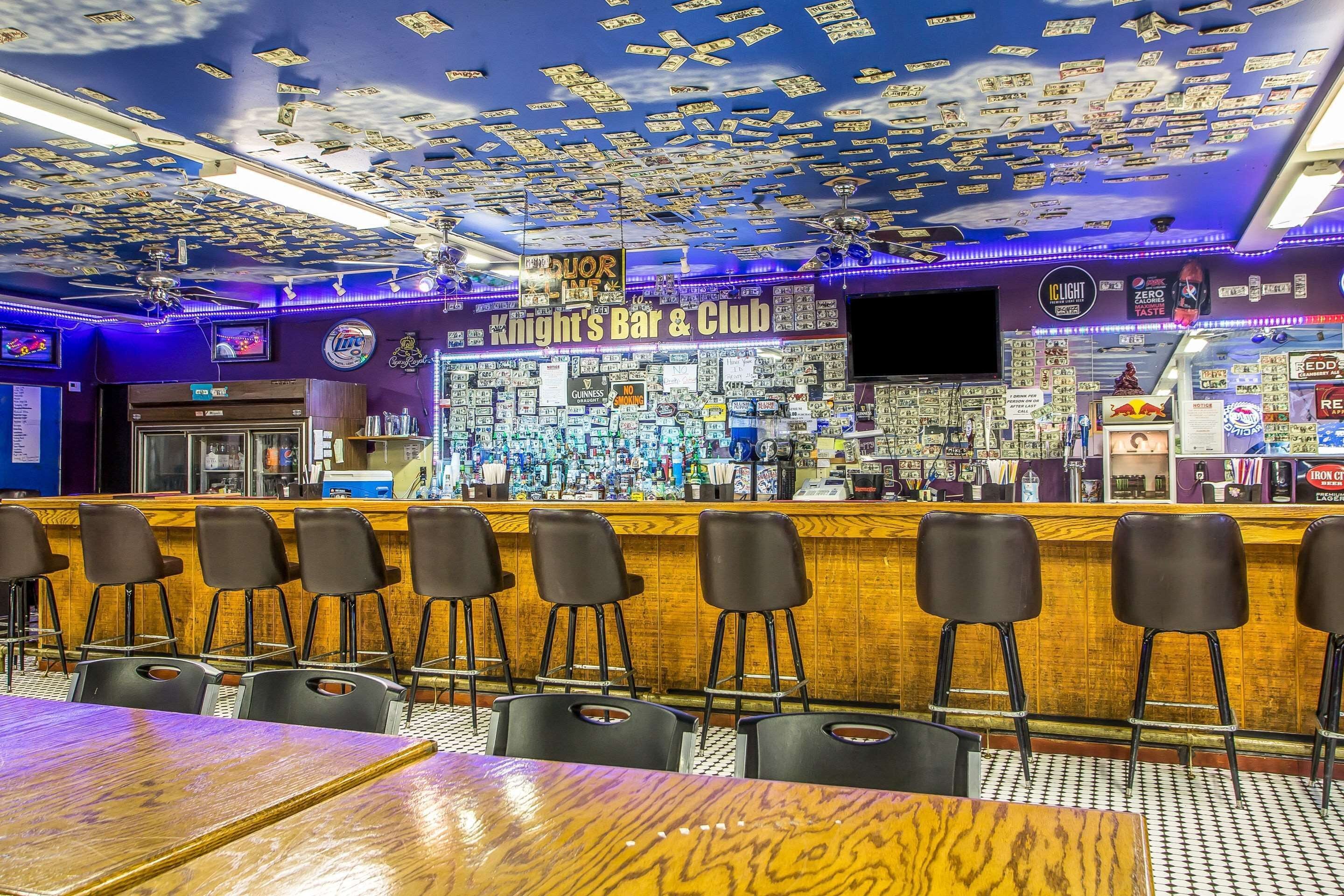 A bar with a ceiling covered in dollar bills, bar stools, and a counter with various bottles and a TV screen.