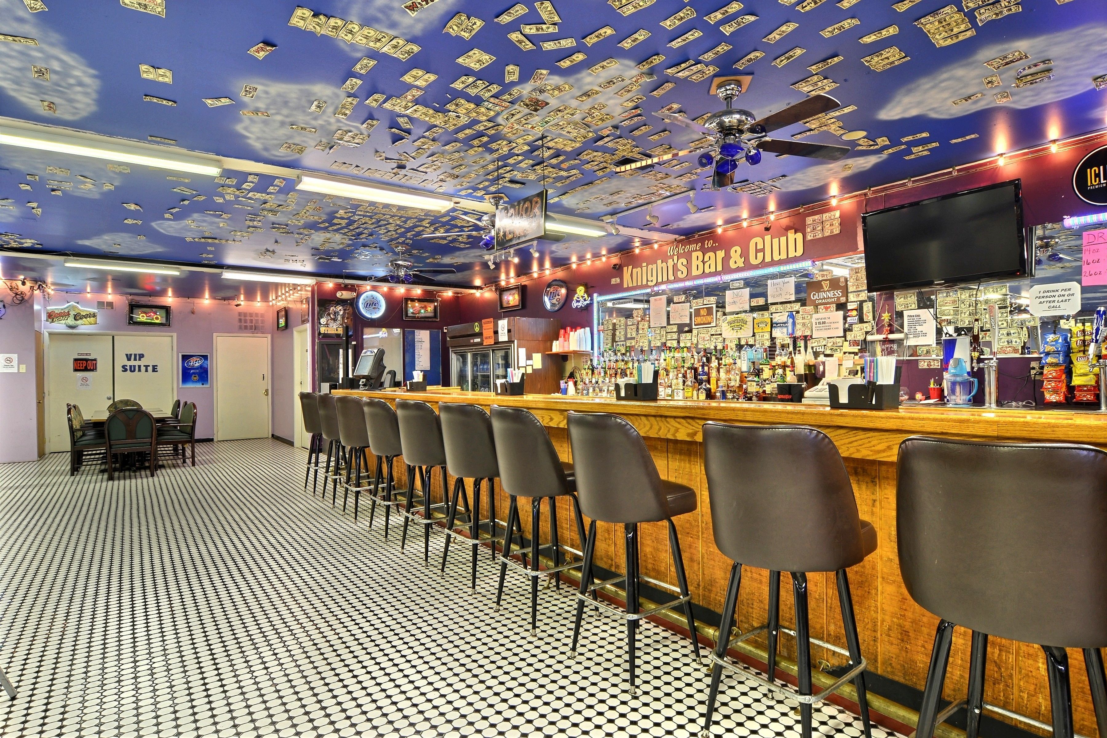 A bar with a ceiling decorated with dollar bills, a bar counter with stools, and a sign reading "Knight's Bar & Club".
