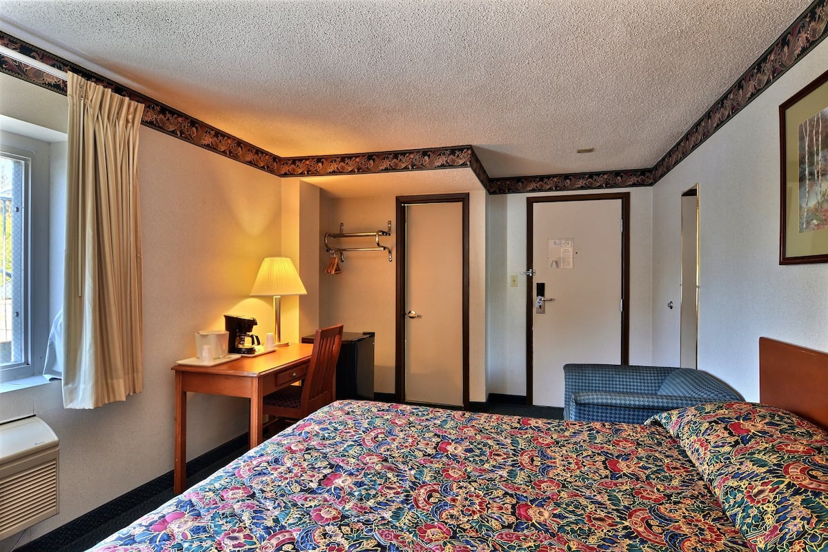 A hotel room with a bed, a desk with a lamp, a chair, a window with curtains, and a door.