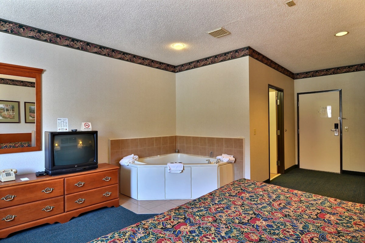 A hotel room with a large bed, a white bathtub, a wooden dresser, a television, and a door.