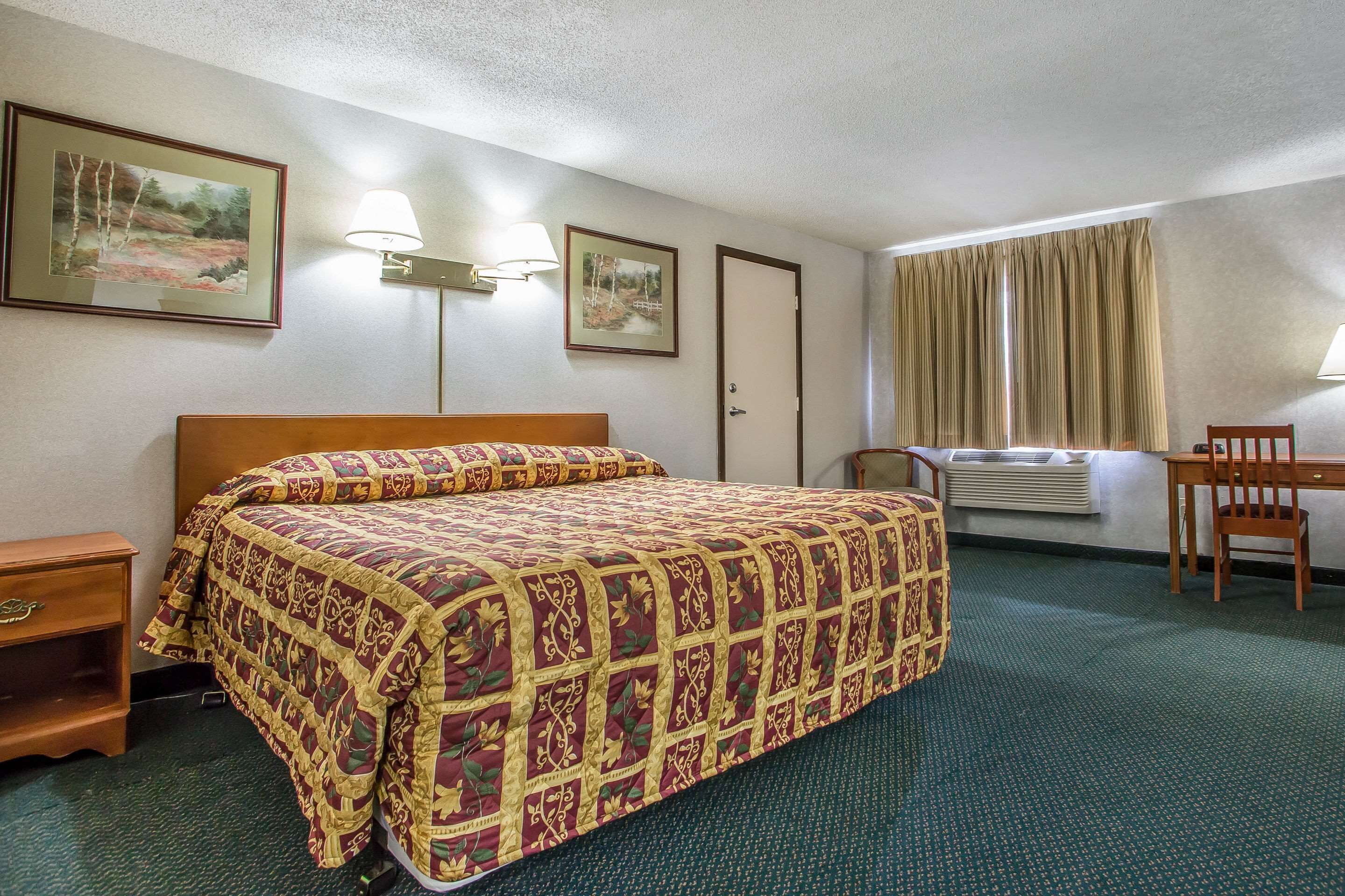 Hotellihuone, jossa on suuri sänky, työpöytä, tuoli, kaksi seinälle asennettua lamppua ja kaksi kehystettyä kuvaa seinällä.