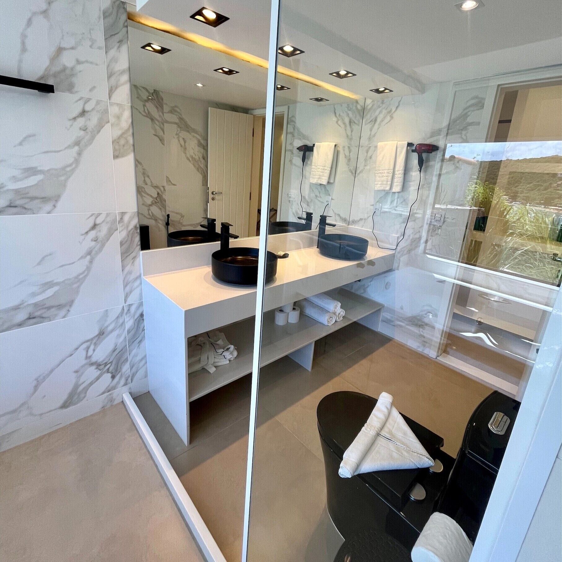 Un baño moderno con paredes de mármol, un espejo grande, dos lavamanos y un inodoro negro.