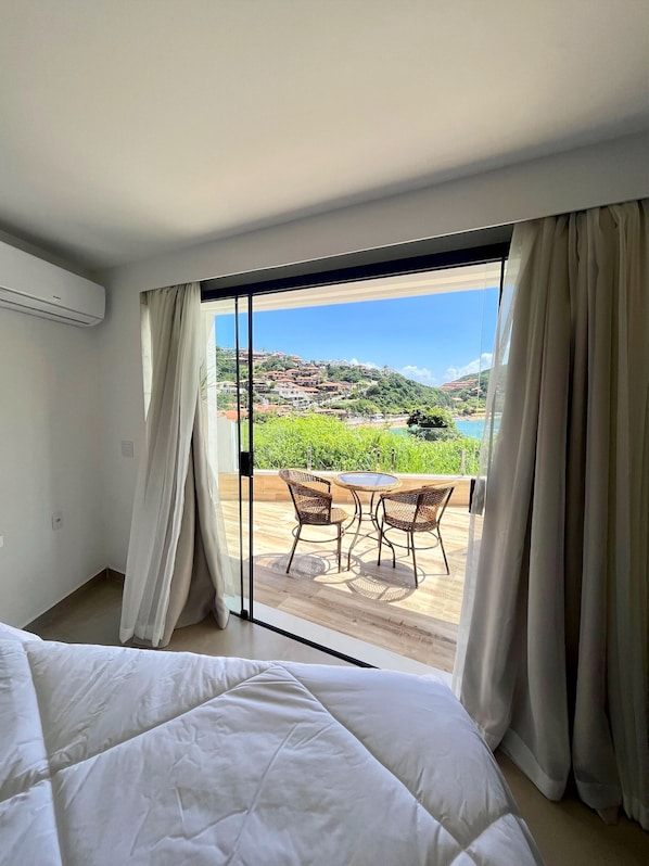 A bedroom with a large bed, a sliding glass door leading to an outdoor area with a table and chairs, and a view of a hillside and water. - Colonna Park Hotel (Búzios)