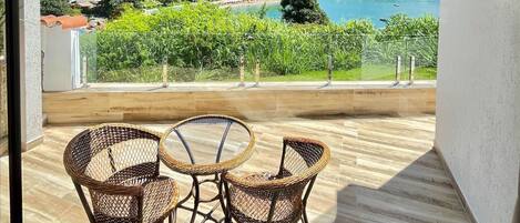 Un balcón con sillas de mimbre y una mesa, con vista a un pueblo costero con casas y una playa.