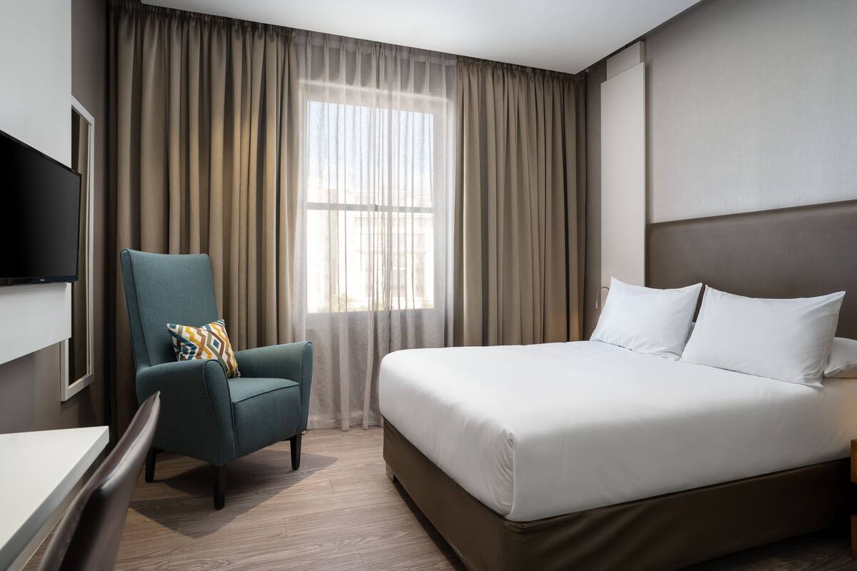 A hotel room with a bed, a chair, a TV, and a window with curtains.