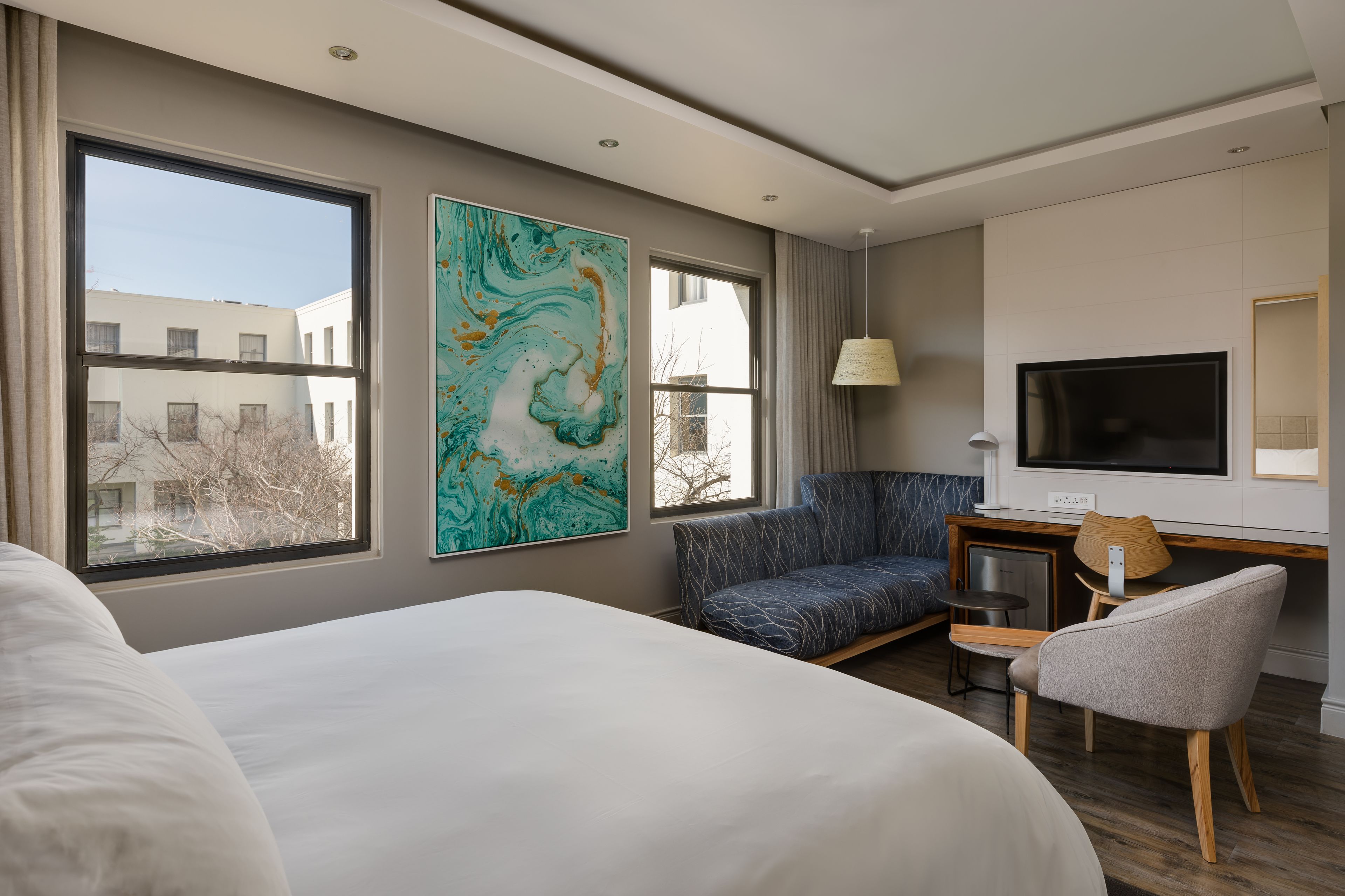 Ein Hotelzimmer mit einem großen Bett, einem Schreibtisch mit Stuhl, einem Fernseher und einem großen Fenster mit Blick auf Gebäude.