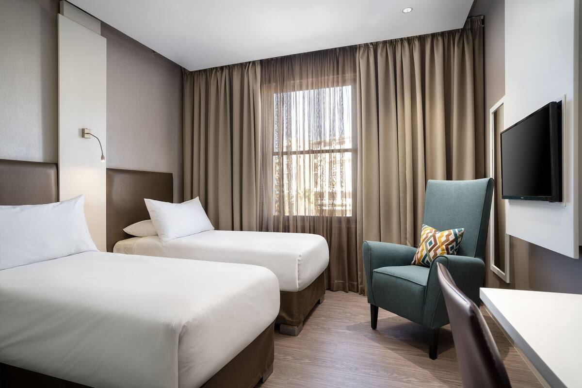 A hotel room with two beds, a desk, a chair, a TV, and a large window with curtains.