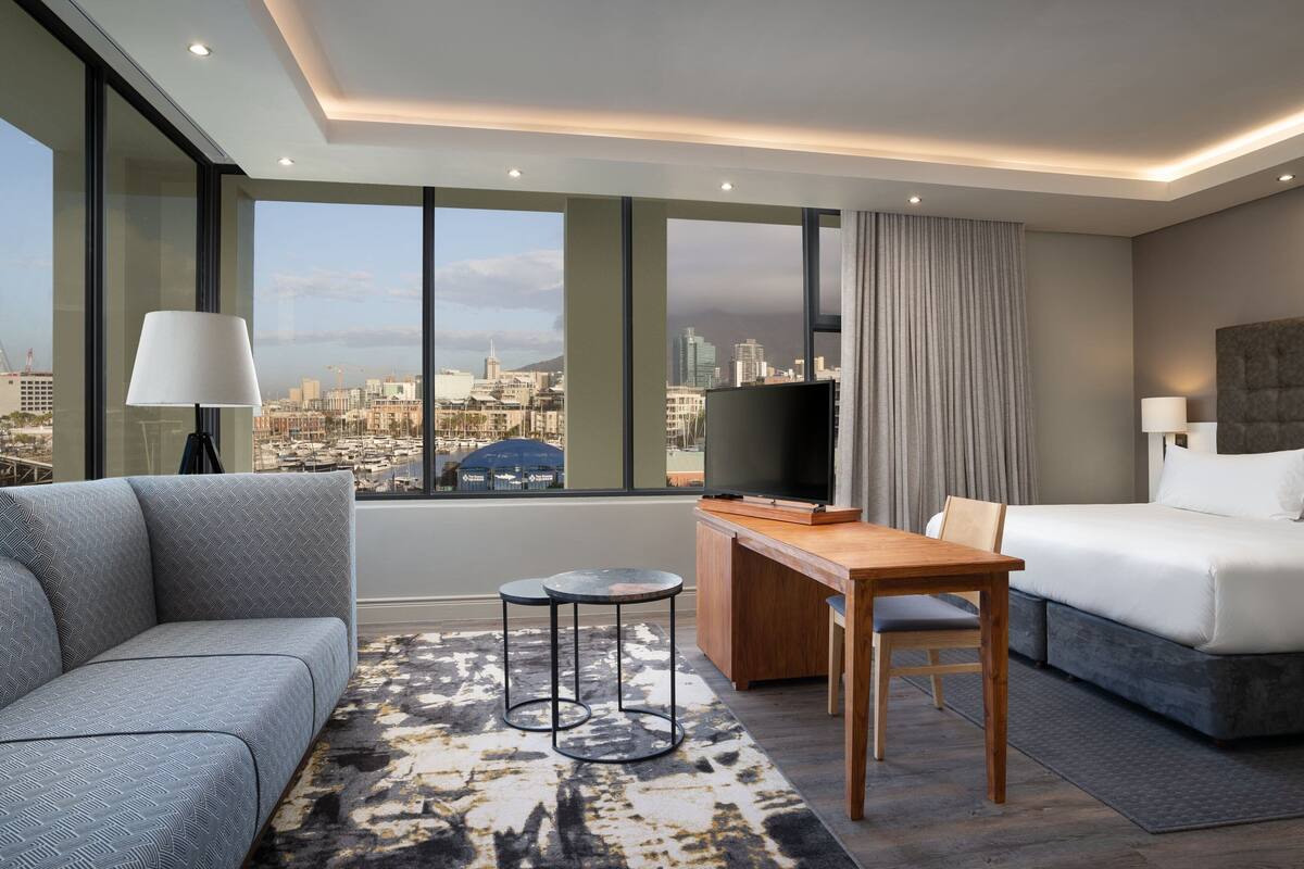 A modern hotel room with a city view, a sofa, a bed, a desk, and a TV.