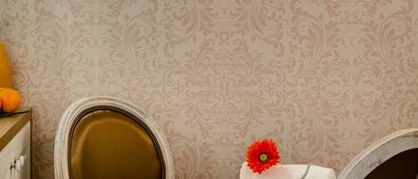 Une salle de bain avec un meuble-lavabo blanc, un miroir et une serviette ornée d’une fleur rouge. On y trouve une étagère avec des articles de toilette et un verre de jus d’orange.