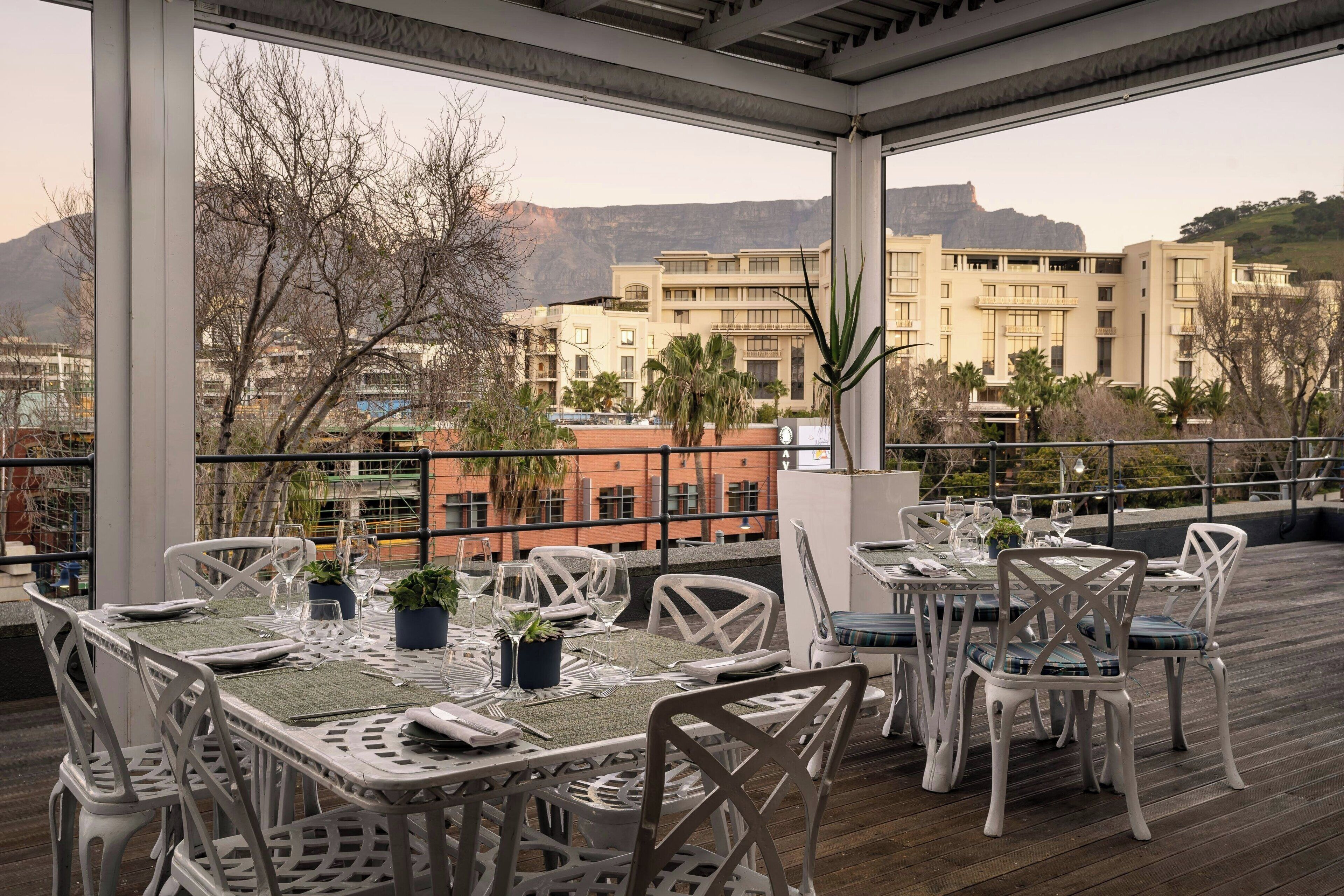 Un espace de restauration en plein air avec des chaises et des tables en métal blanc, le tout mis en valeur par un décor composé d’immeubles et de montagnes.