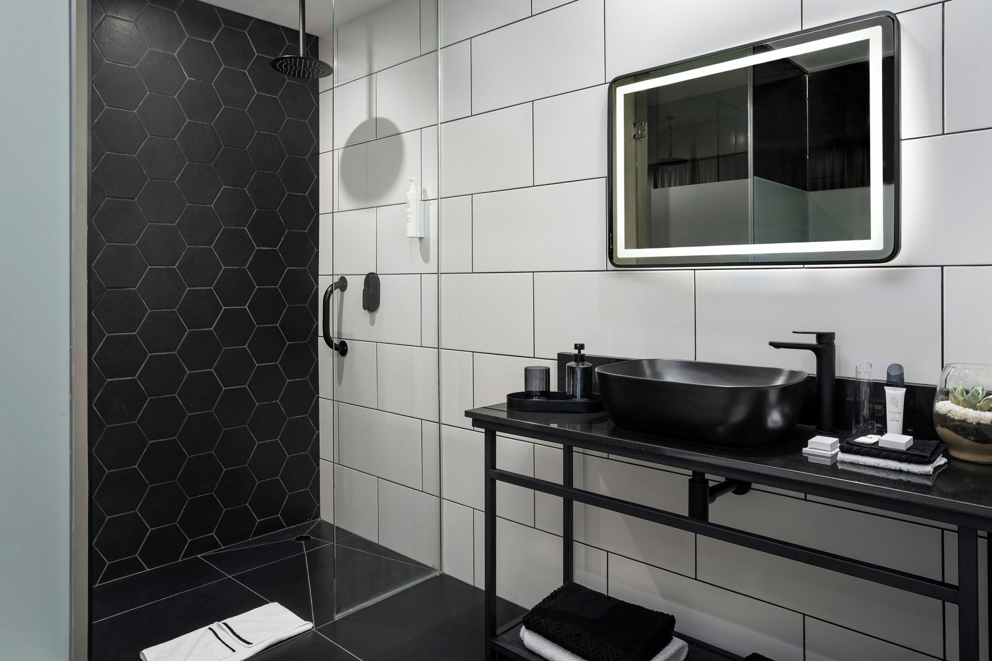 Een moderne badkamer met een douchewand betegeld met zwarte honingraatmozaïek, een zwart wastafelmeubel met een zwart keramiekbakje en een spiegel met verlichting rondom.