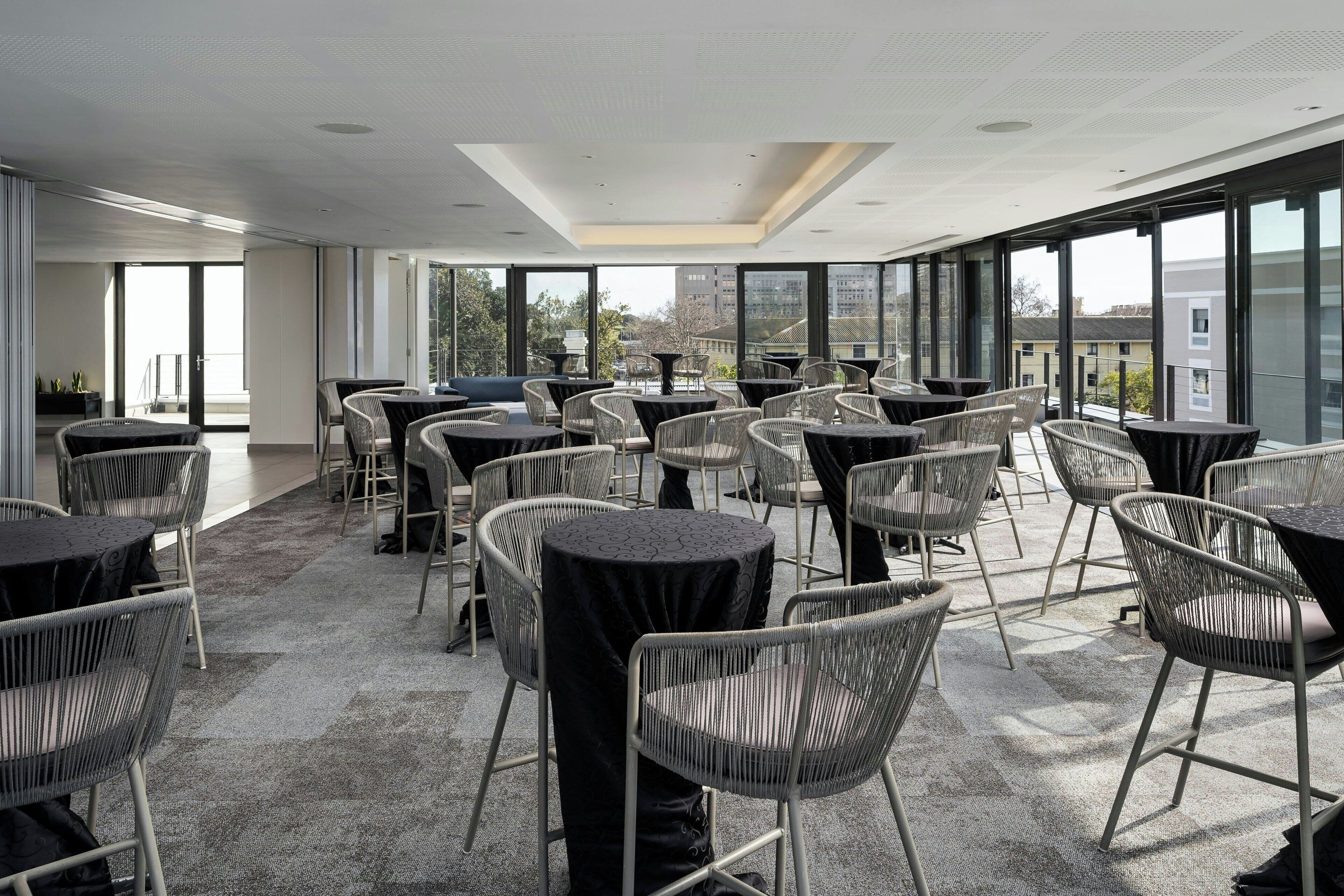 A modern indoor dining area with round tables and wicker chairs, large windows offering views of the city.