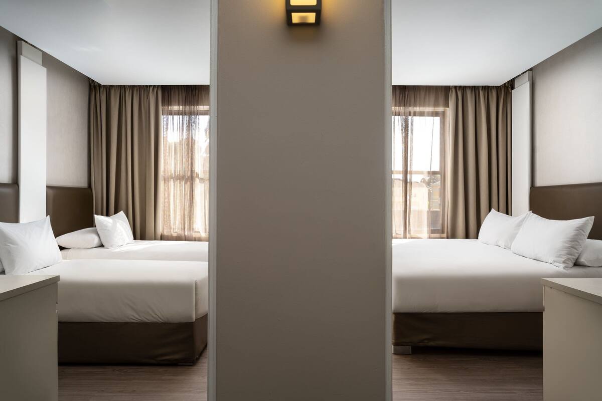 A hotel room with two beds, a nightstand, a wall-mounted light, and a window with curtains.