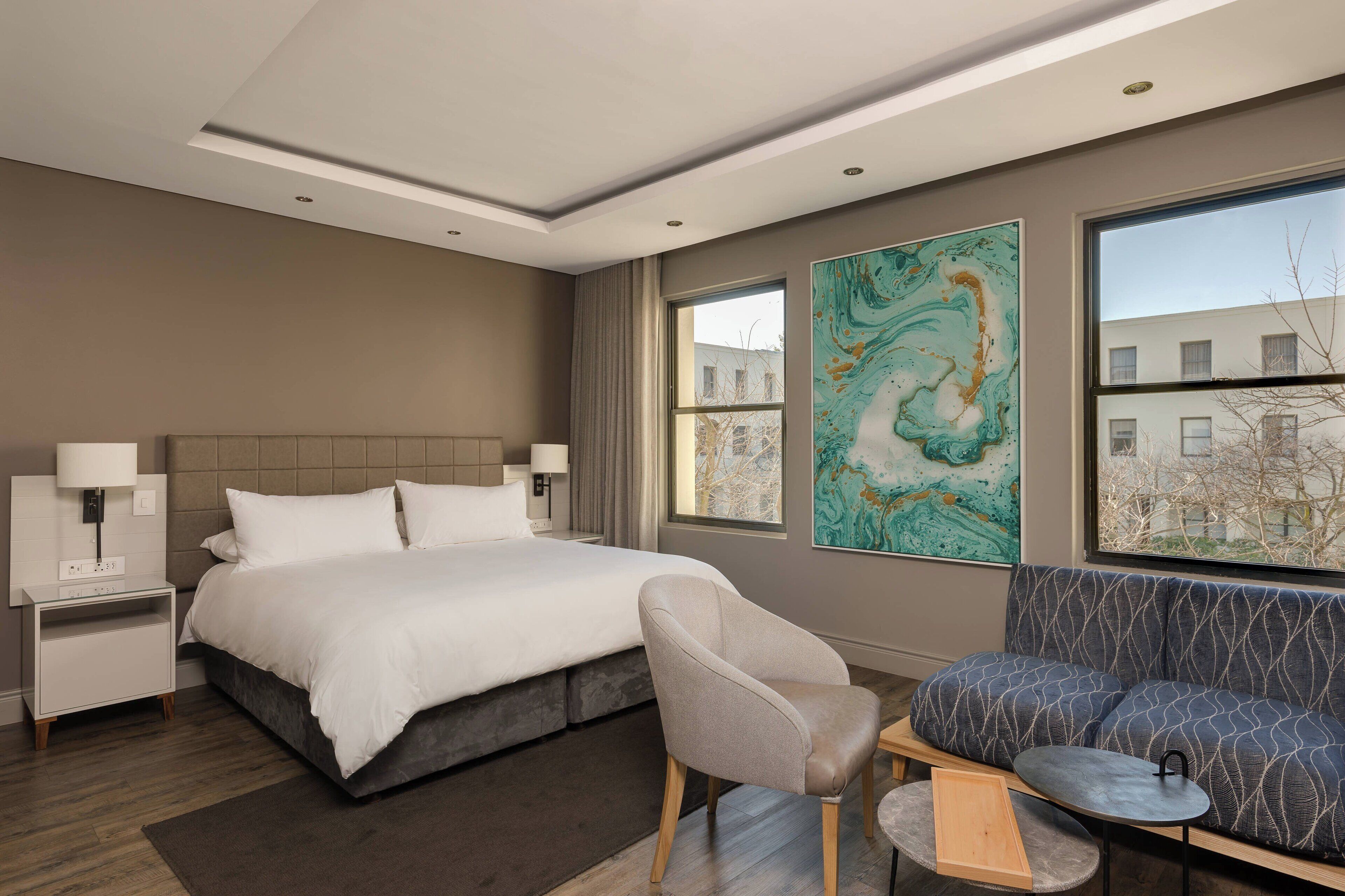Una habitación de hotel moderna con una cama grande, zona de estar y un amplio ventanal con vistas a edificios.
