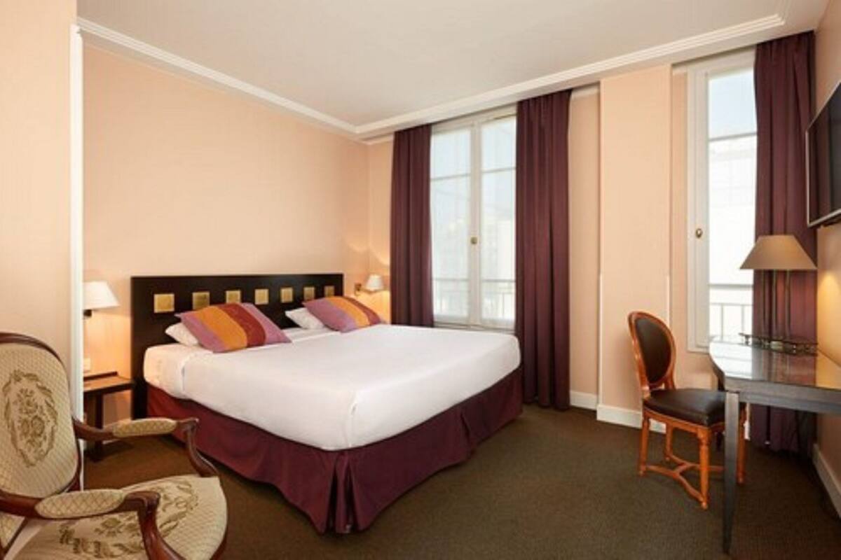 A hotel room with a large bed, a desk, a chair, a TV, and a window with curtains.