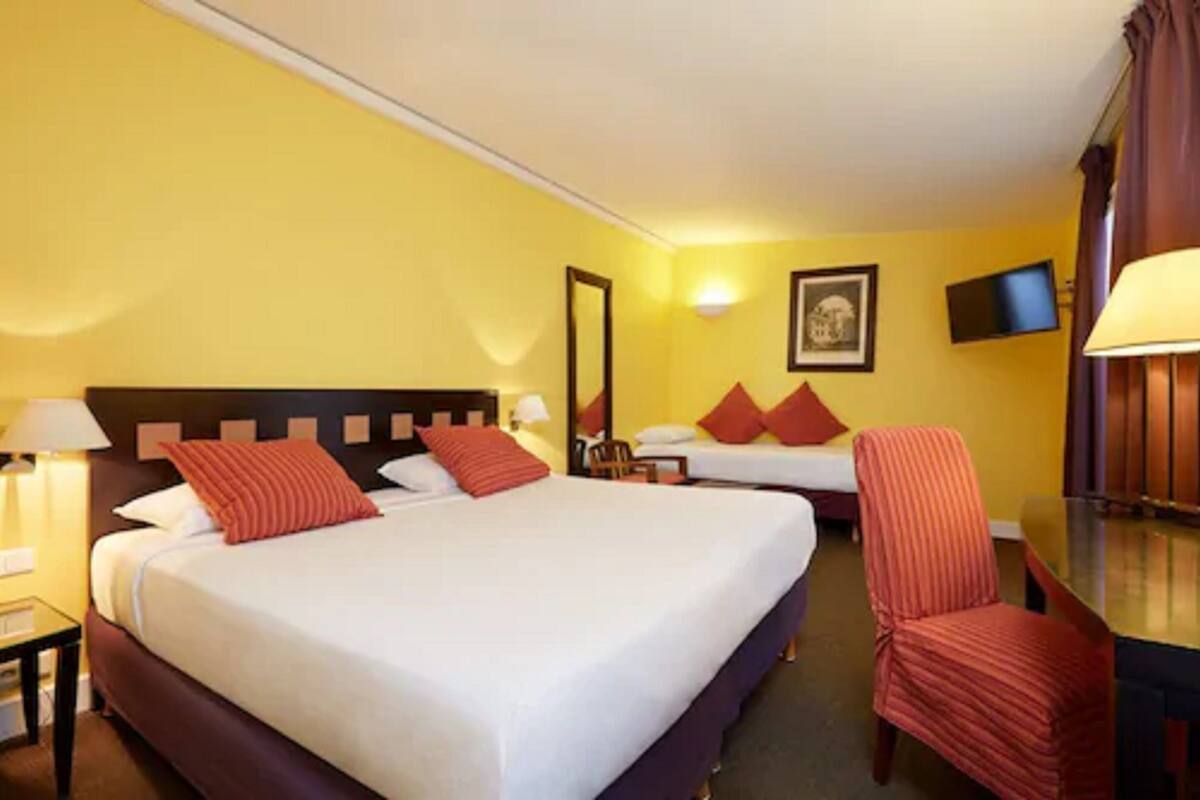 A hotel room with two beds, a desk, a chair, a TV, and a lamp.