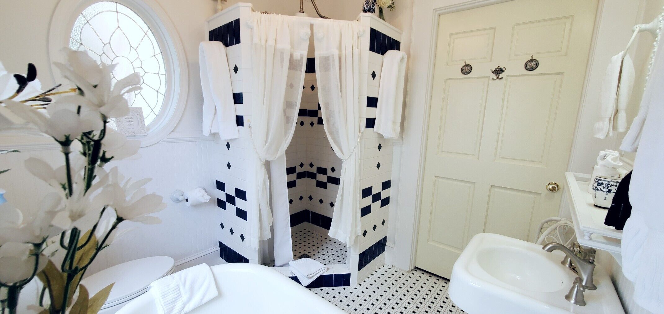 Un baño con un lavamanos blanco, un inodoro blanco, una bañera blanca, una ducha blanca con diseño de cuadros y una puerta blanca con ventana.