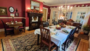 A dining room with a large table set for six, surrounded by chairs. A fireplace with a mantle and a mirror above it is visible. The room has red walls and a patterned rug on the floor. - Pettigru Place Bed & Breakfast (Greenville)