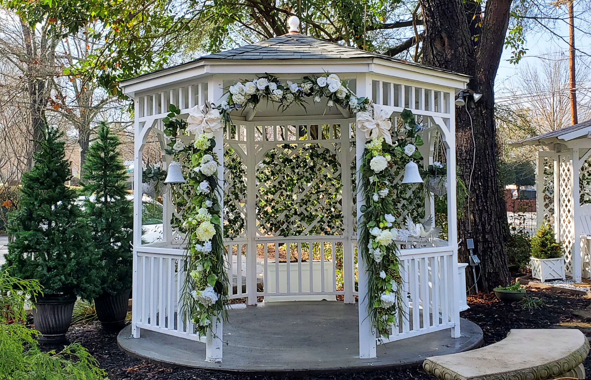 Un belvédère aux balustrades blanches et aux décorations florales, entouré d’arbres et de plantes en pot.